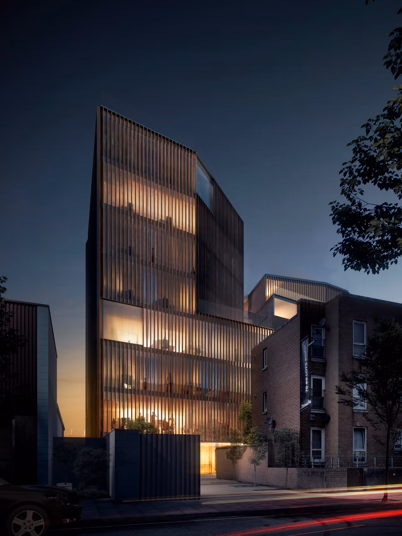 78-89 Lots Road in Chelsea, London at dusk, illuminated glass and timber-louvred office building beside a brick residential street, warm interior lighting glowing through