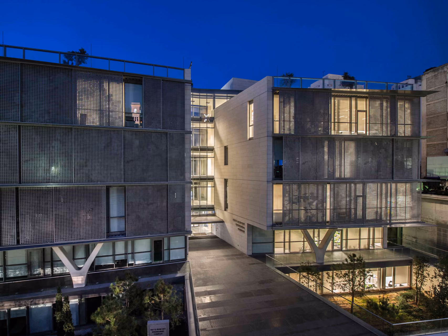 Night view of AUB IOEC Engineering Labs in Beirut, twin perforated-metal and stone volumes flanking a lit atrium bridge, illuminated interiors visible through glazing