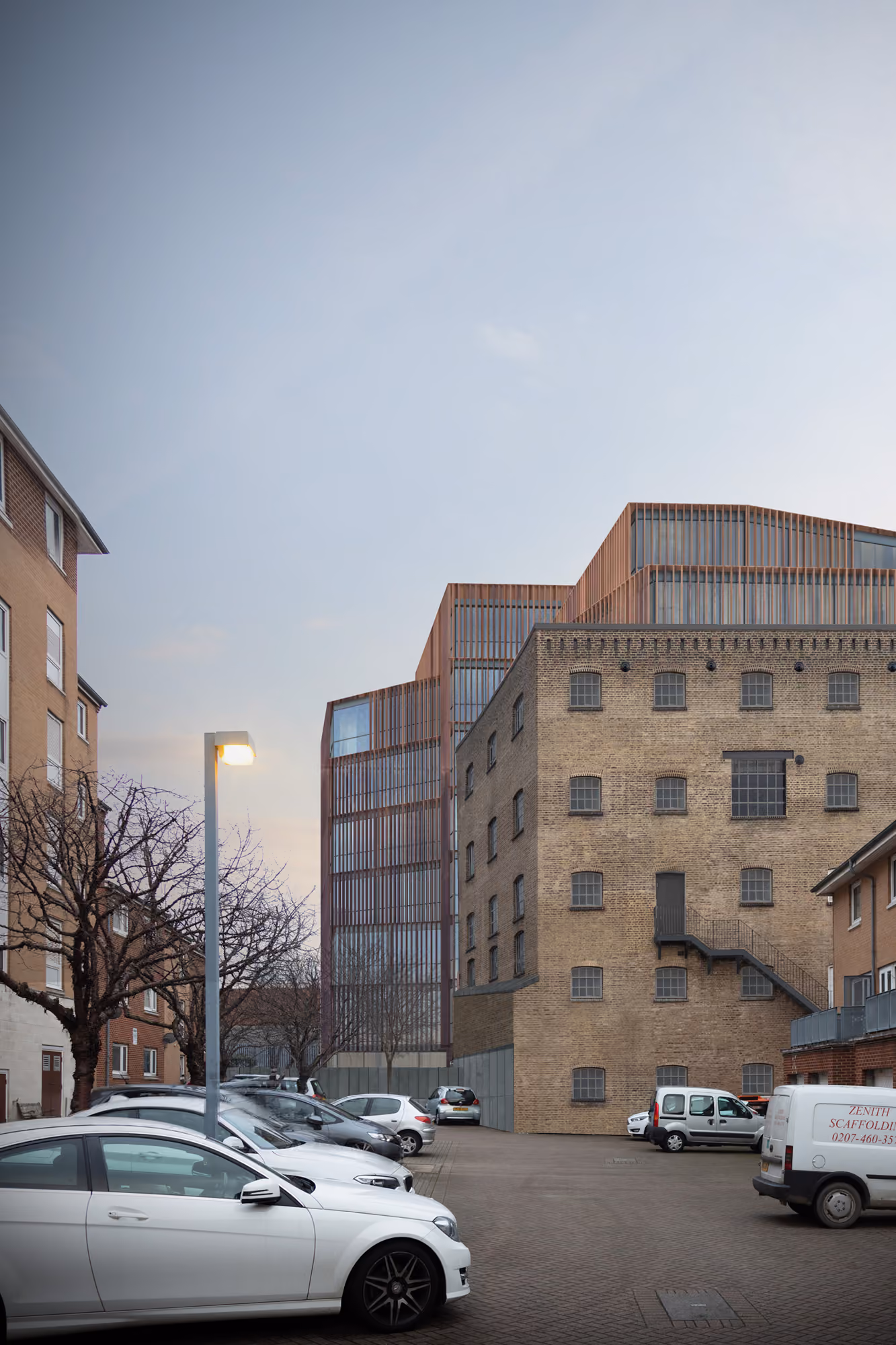 78-89 Lots Road in Chelsea, London, new copper-toned upper floors rising behind a retained brick industrial building, seen from a car park at dusk