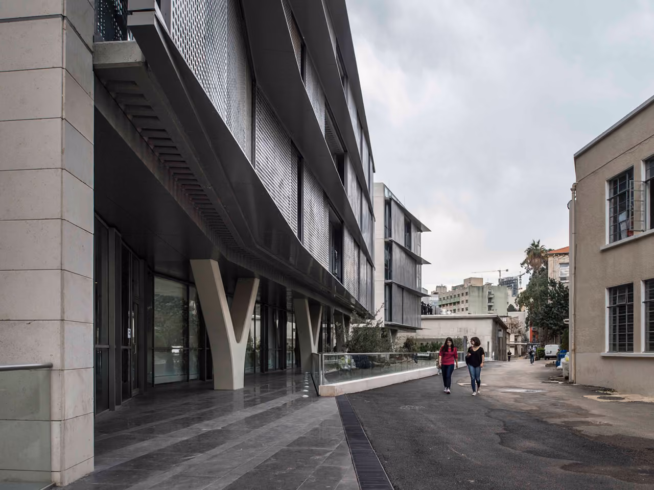 Perforated metal-clad facade of the AUB IOEC Engineering Labs in Beirut, cantilevered on V-shaped columns above a sheltered walkway with pedestrians