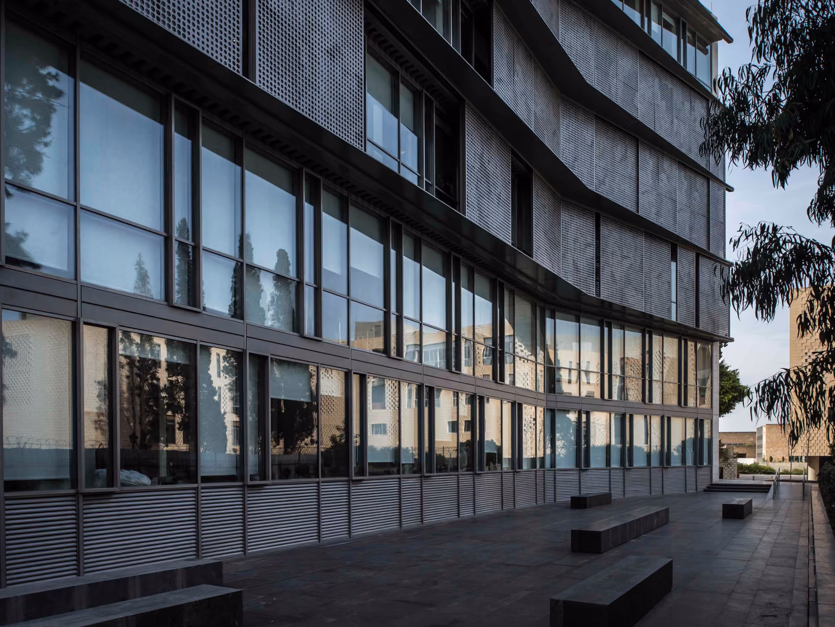 Curved glass facade of the AUB IOEC Engineering Labs in Beirut, perforated metal sunshades above and a paved courtyard with linear stone benches below