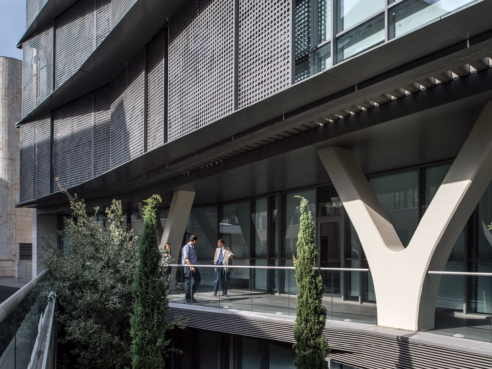 Perforated metal facade and Y-shaped columns at AUB IOEC Engineering Labs in Beirut, with people conversing on a plant-lined balcony walkway