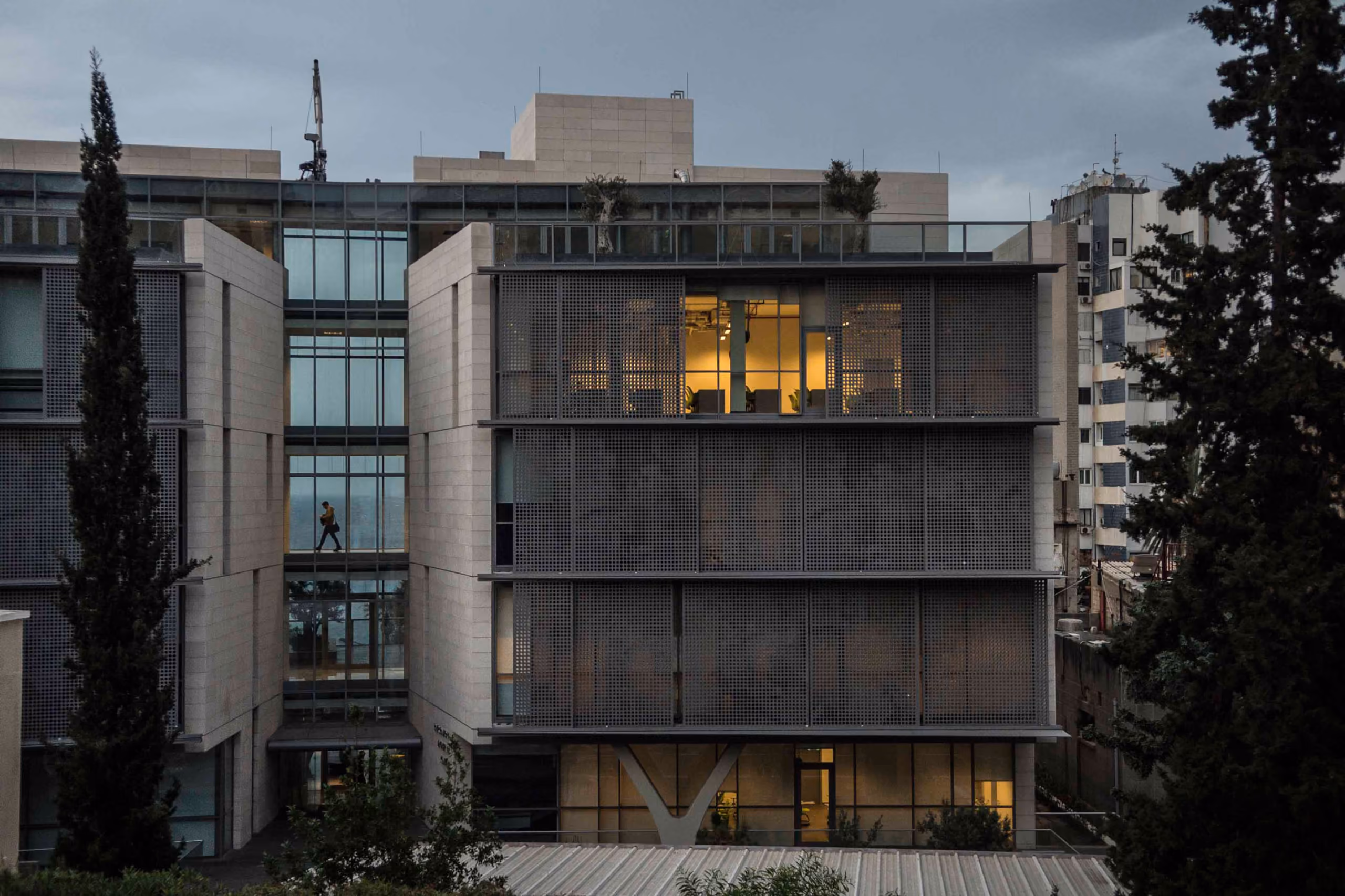 Perforated metal screens shade the glazed facade of AUB IOEC Engineering Labs in Beirut, with lit interiors visible at dusk between stone volumes