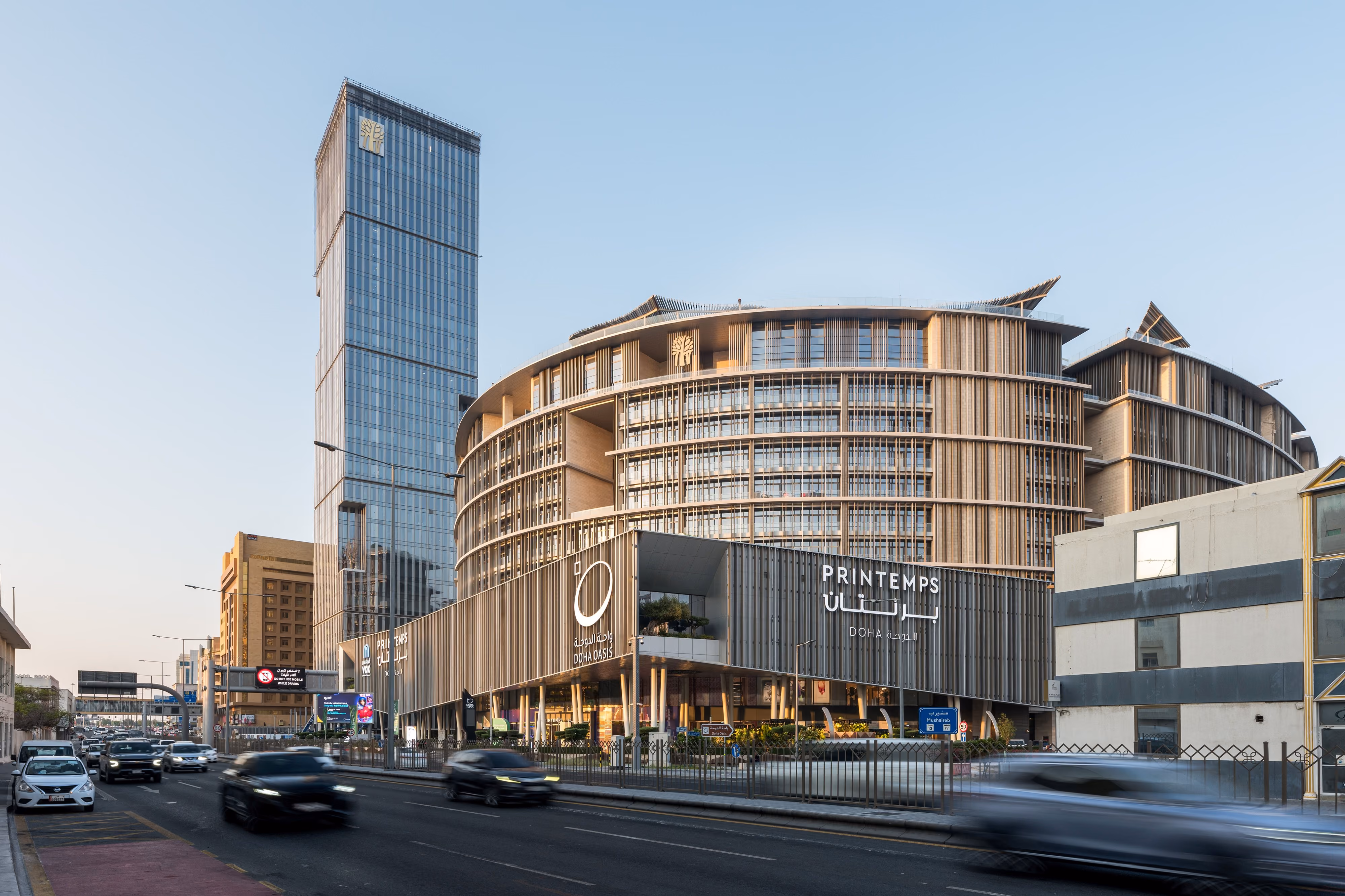 Doha Oasis, a mixed-use complex by Nabil Gholam Architects with curved timber-louvered facades and a slender glass tower, at street level in Doha, Qatar