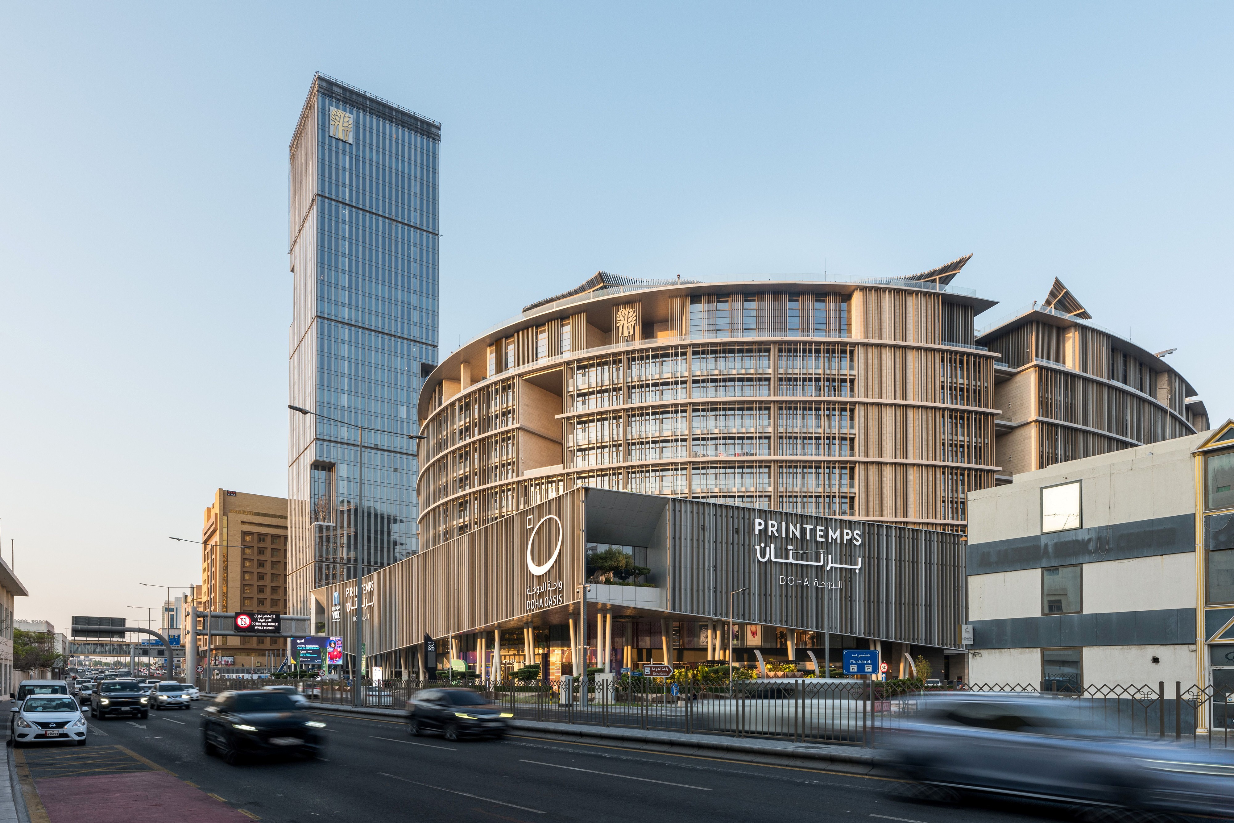 Doha Oasis, a mixed-use complex by Nabil Gholam Architects with curved timber-louvered facades and a slender glass tower, at street level in Doha, Qatar
