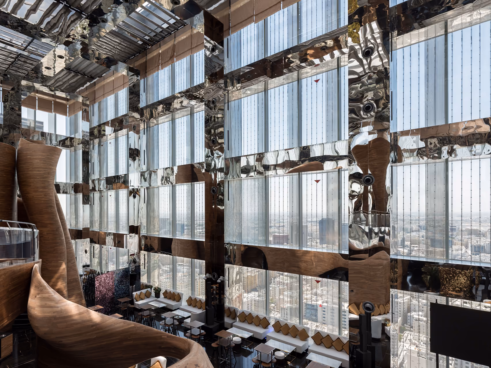 High-level lounge in Doha Oasis, curved wooden staircase beside a mirrored double-height wall with sweeping views over the city skyline
