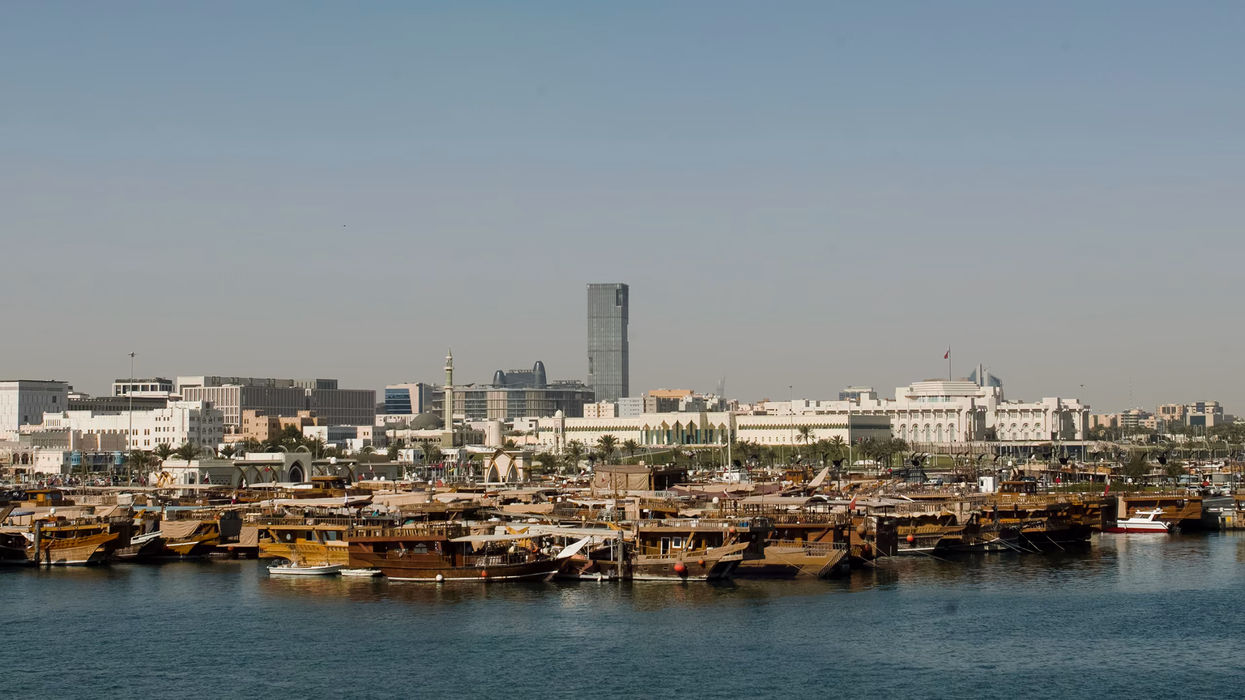 Doha Oasis site context in Doha, Qatar, traditional dhows moored along the waterfront with the city skyline and a lone tower behind