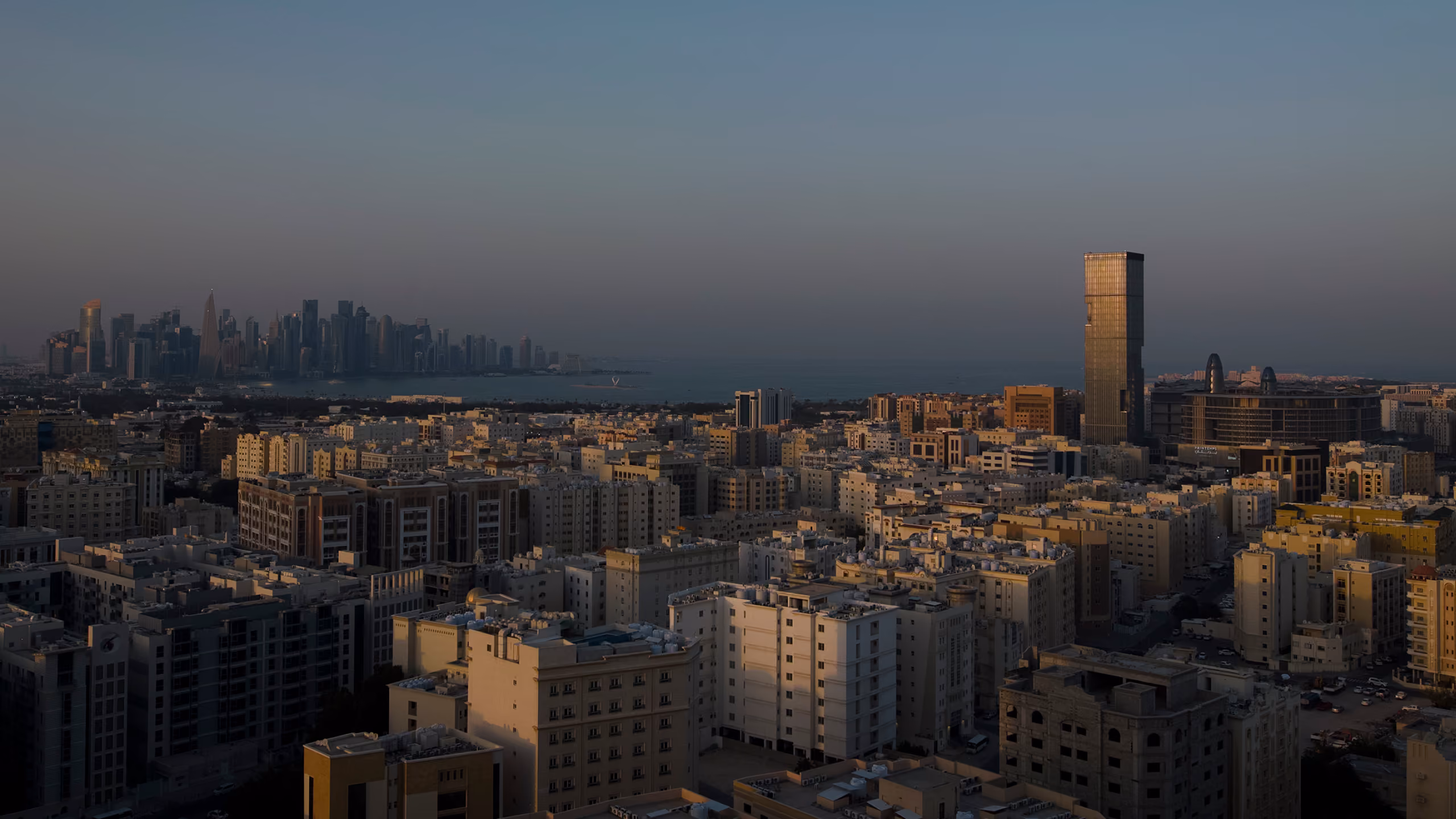 Aerial dusk view of Doha Oasis's slender tower rising above the low-rise cityscape, with Doha's skyline and coastline visible in the distance