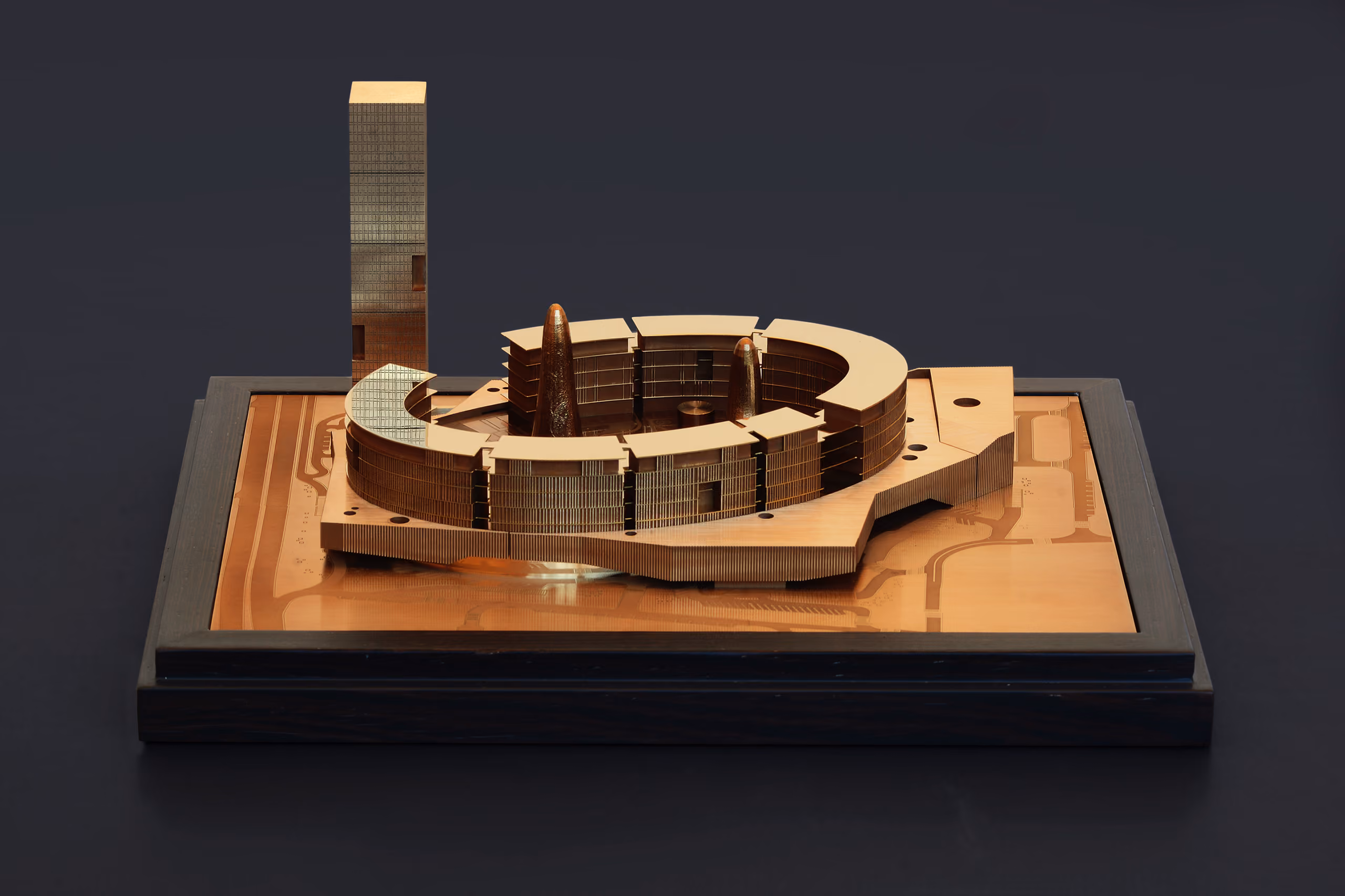 Bronze architectural model of Doha Oasis in Doha, showing a curved ring-shaped podium encircling a courtyard, with a slender tower rising alongside on an engraved site plate
