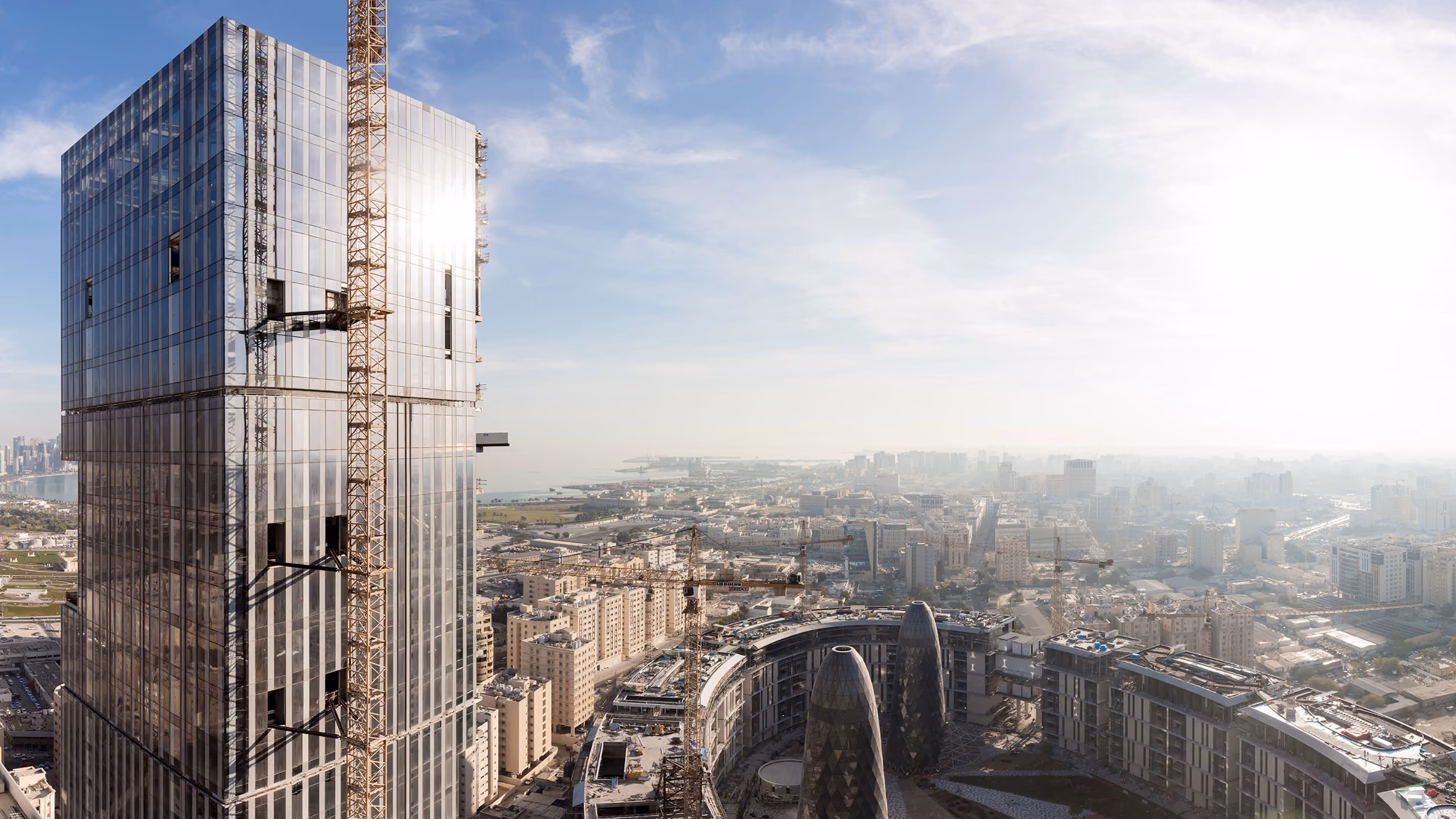 Aerial view of Doha Oasis in Doha under construction, glass tower with crane in foreground overlooking the low-rise complex and city skyline