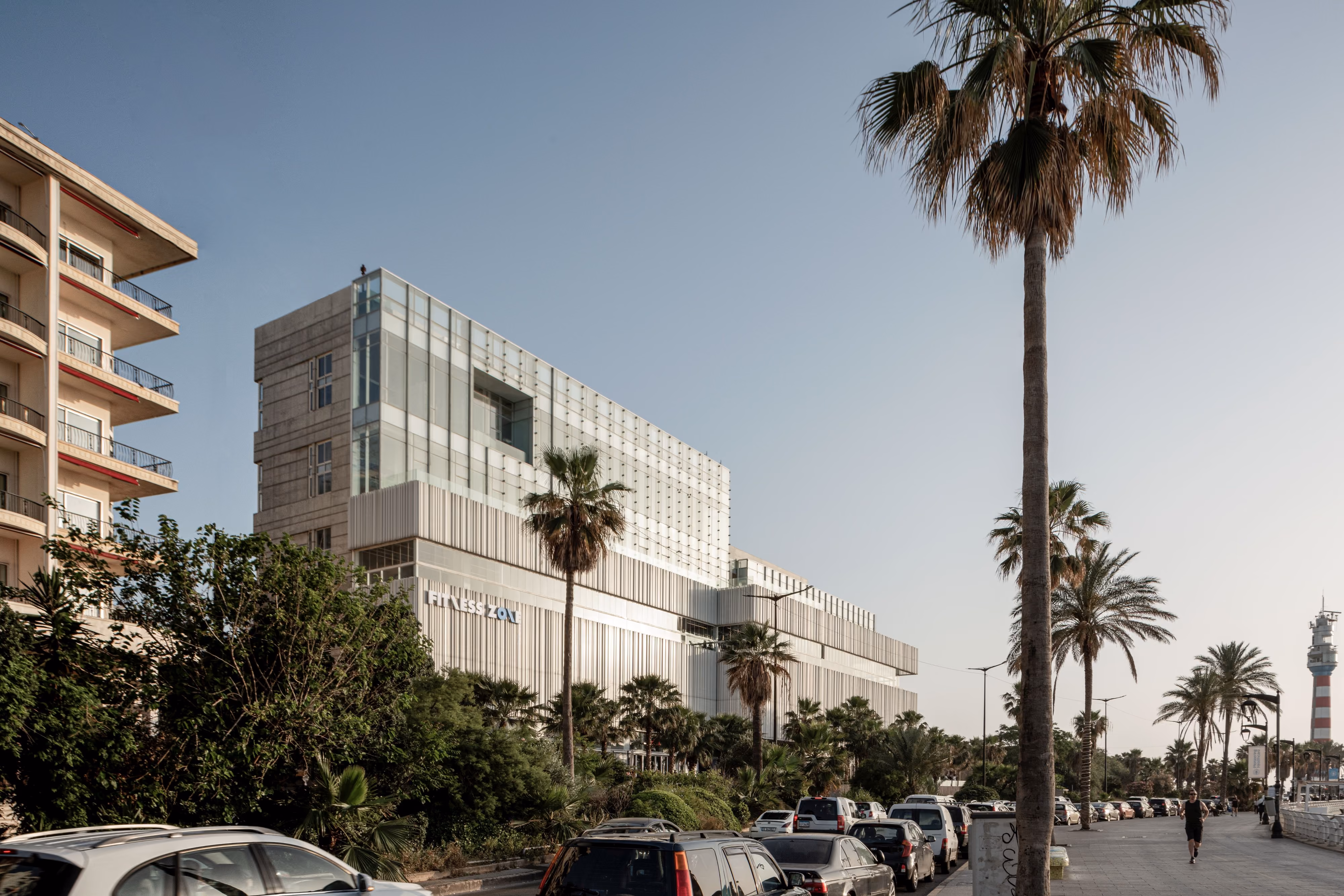 Dalfa Seafront in Beirut, a striped mixed-use volume with a glazed tower above, seen from the palm-lined Corniche with parked cars in the foreground