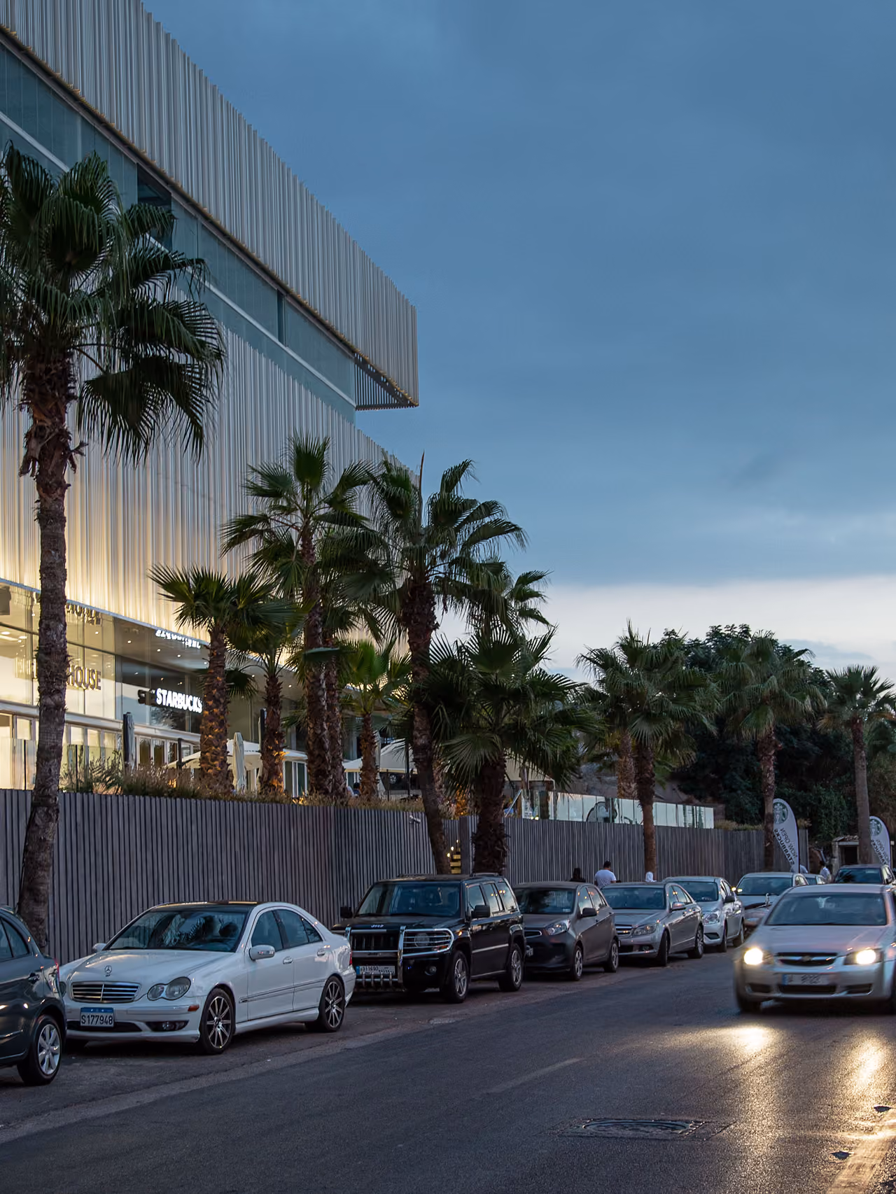 Street-level view of Dalfa Seafront in Beirut at dusk, palm-lined corniche with parked cars beneath the glowing perforated metal facade of the podium