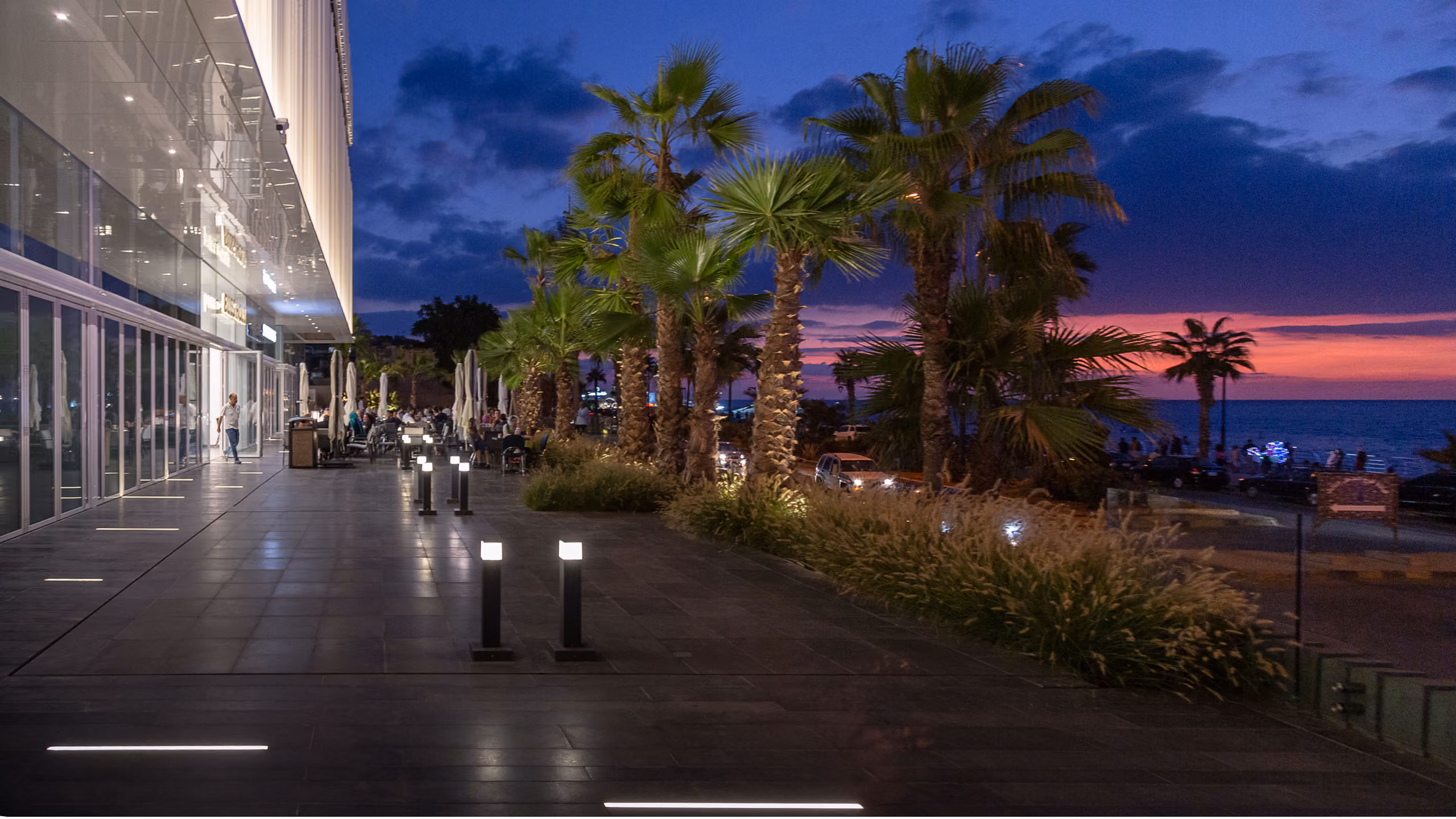 Evening view of Dalfa Seafront in Beirut, illuminated palm-lined promenade beside the glazed podium facade with the sunset over the sea beyond