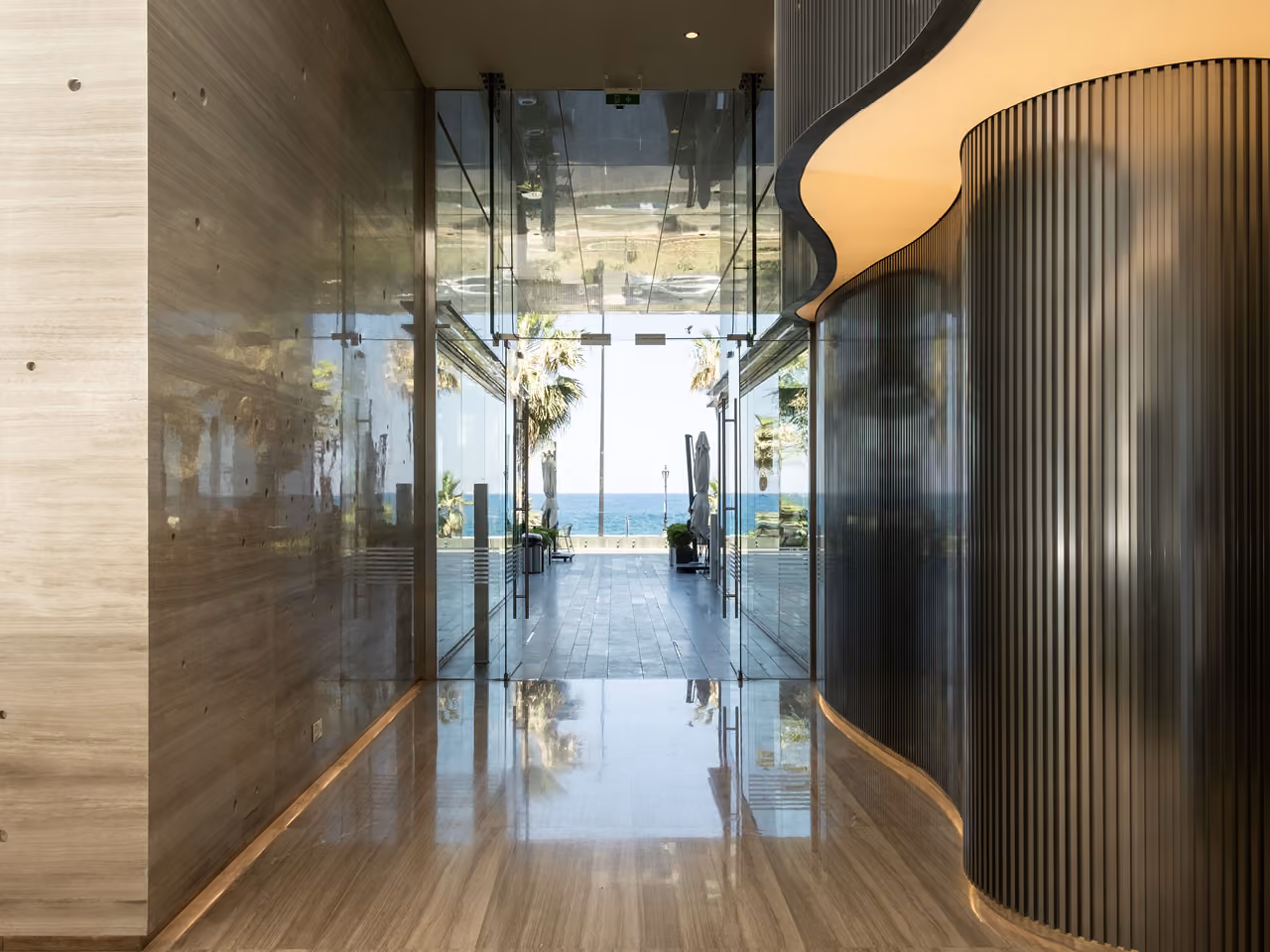 Ground-floor lobby of Dalfa Seafront in Beirut, glass corridor with fluted dark panelling leading to a sea view framed by palm trees