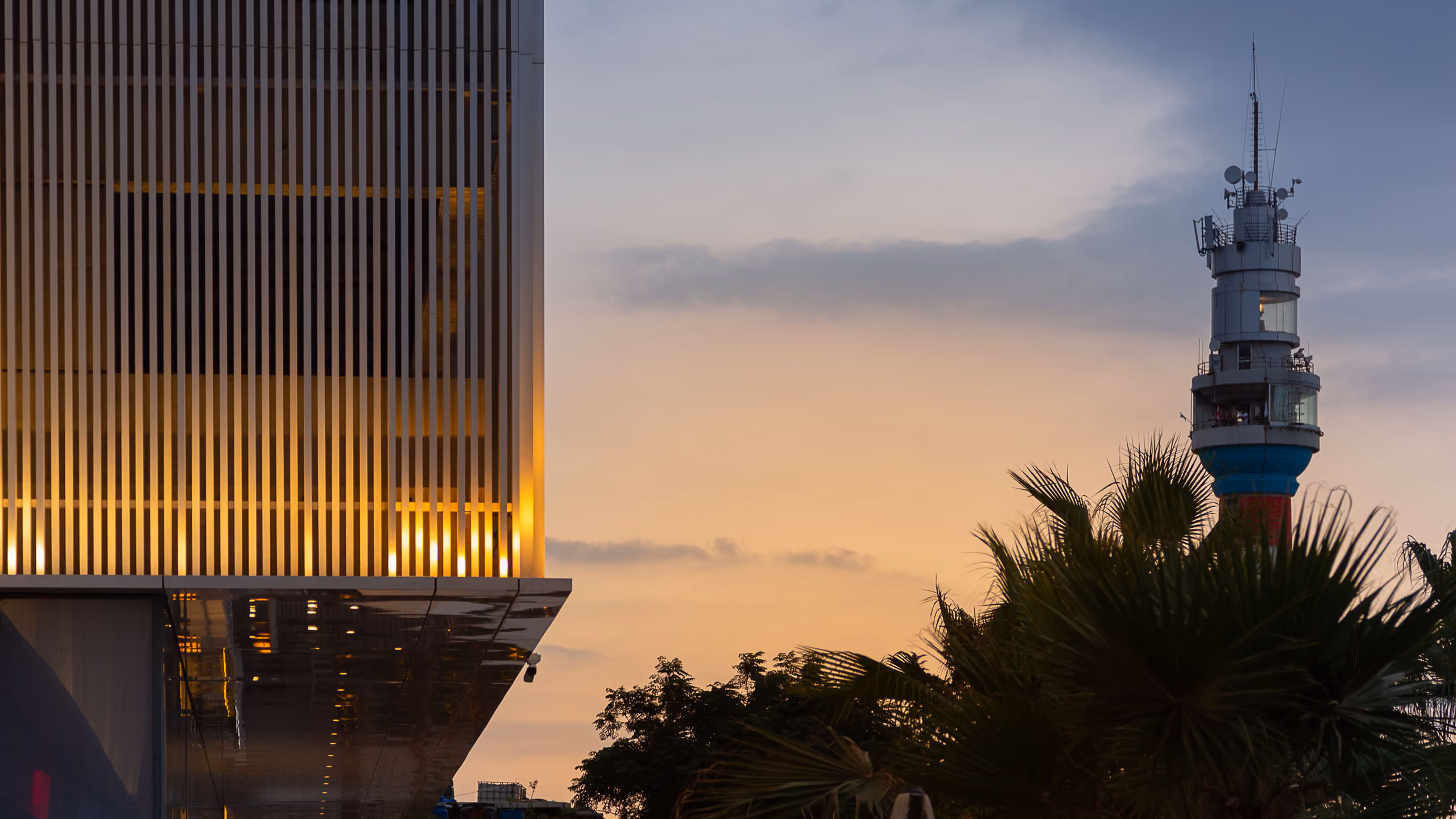 Vertical louvred facade of Dalfa Seafront in Beirut glowing at dusk, with a lighthouse tower and palm trees against an orange sky