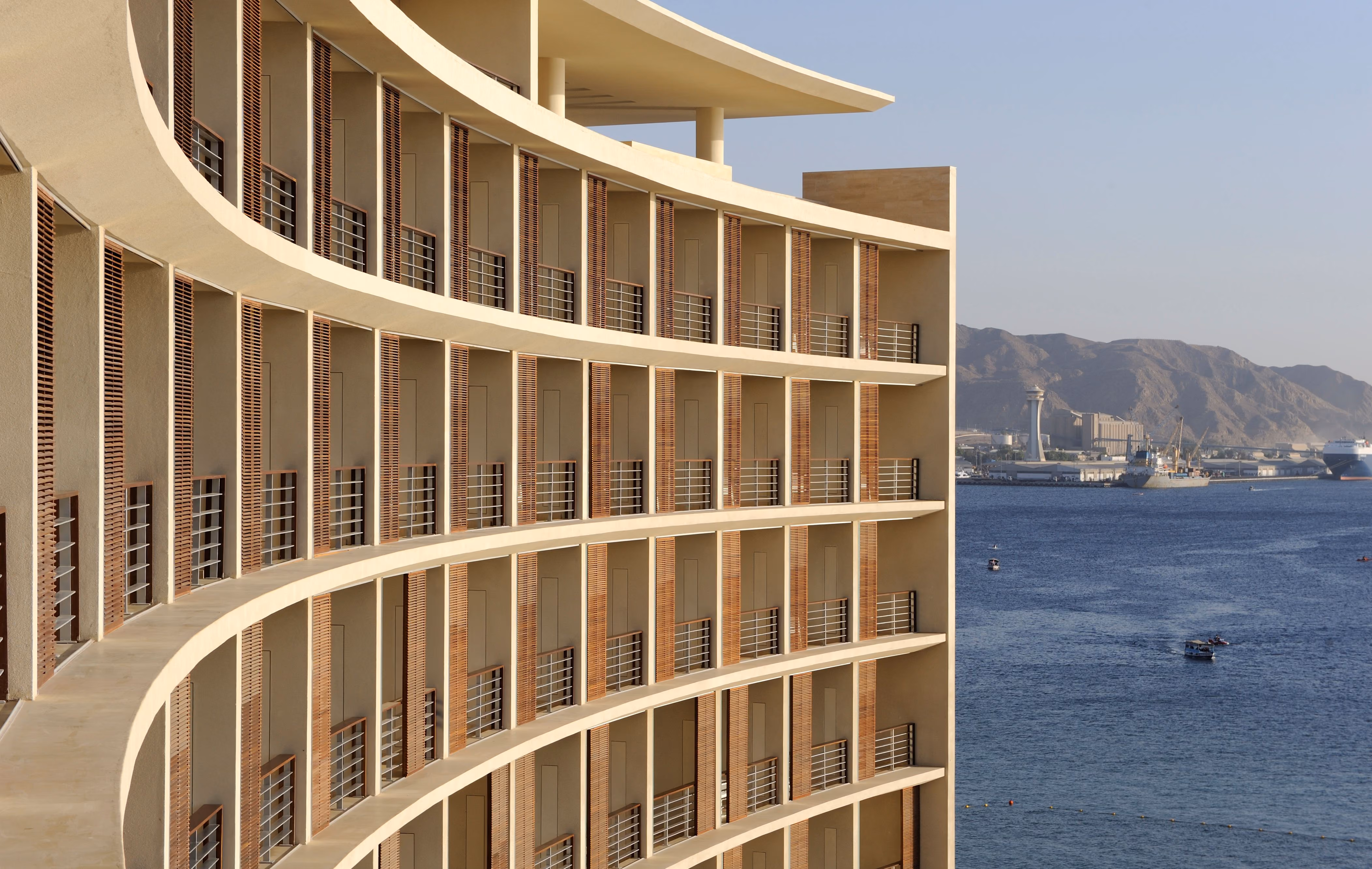 Curved yellow-stone facade of the Kempinski Hotel in Aqaba, tiered balconies with timber louvres overlooking the sea and distant red mountains