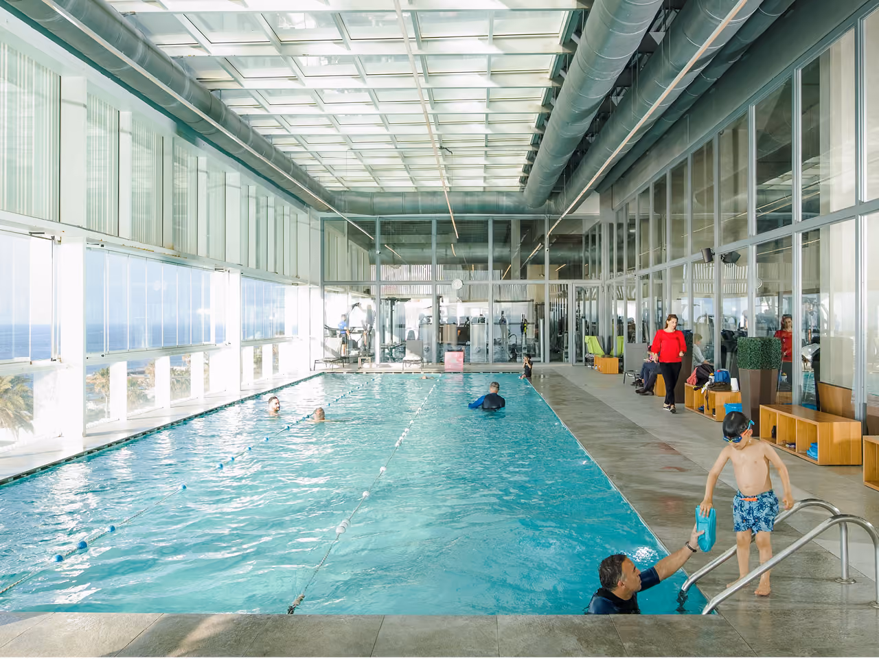 Indoor lap pool at Dalfa Seafront in Beirut, swimmers and a child at the steps, glass walls opening to sea views under an exposed ductwork ceiling