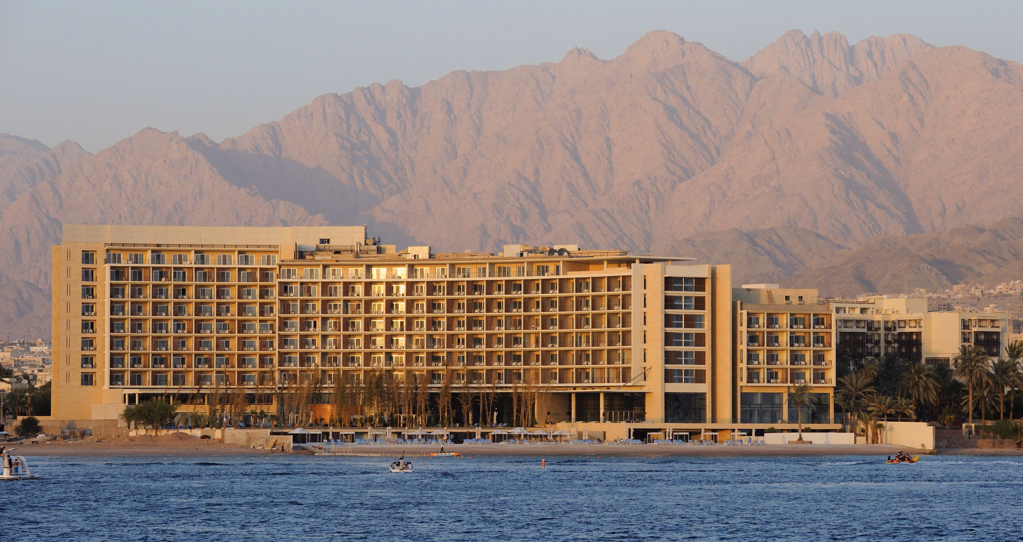 Kempinski Hotel in Aqaba, curved yellow stone facade with tiered balconies along the beach, set against dramatic red mountains and the sea