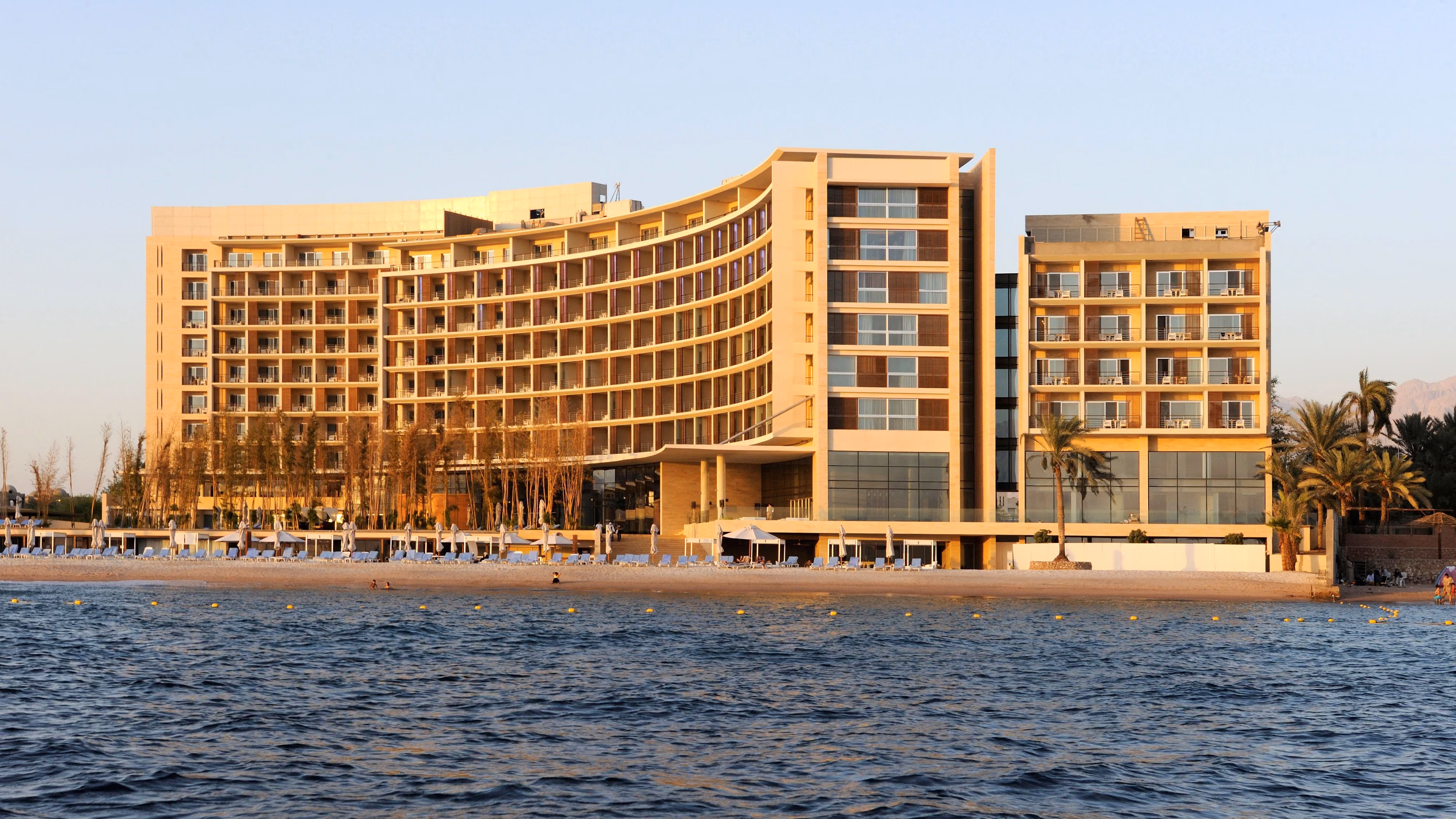 Kempinski Hotel by Nabil Gholam Architects, a curved beachfront hotel in golden stone rising along the shoreline in Aqaba, Jordan