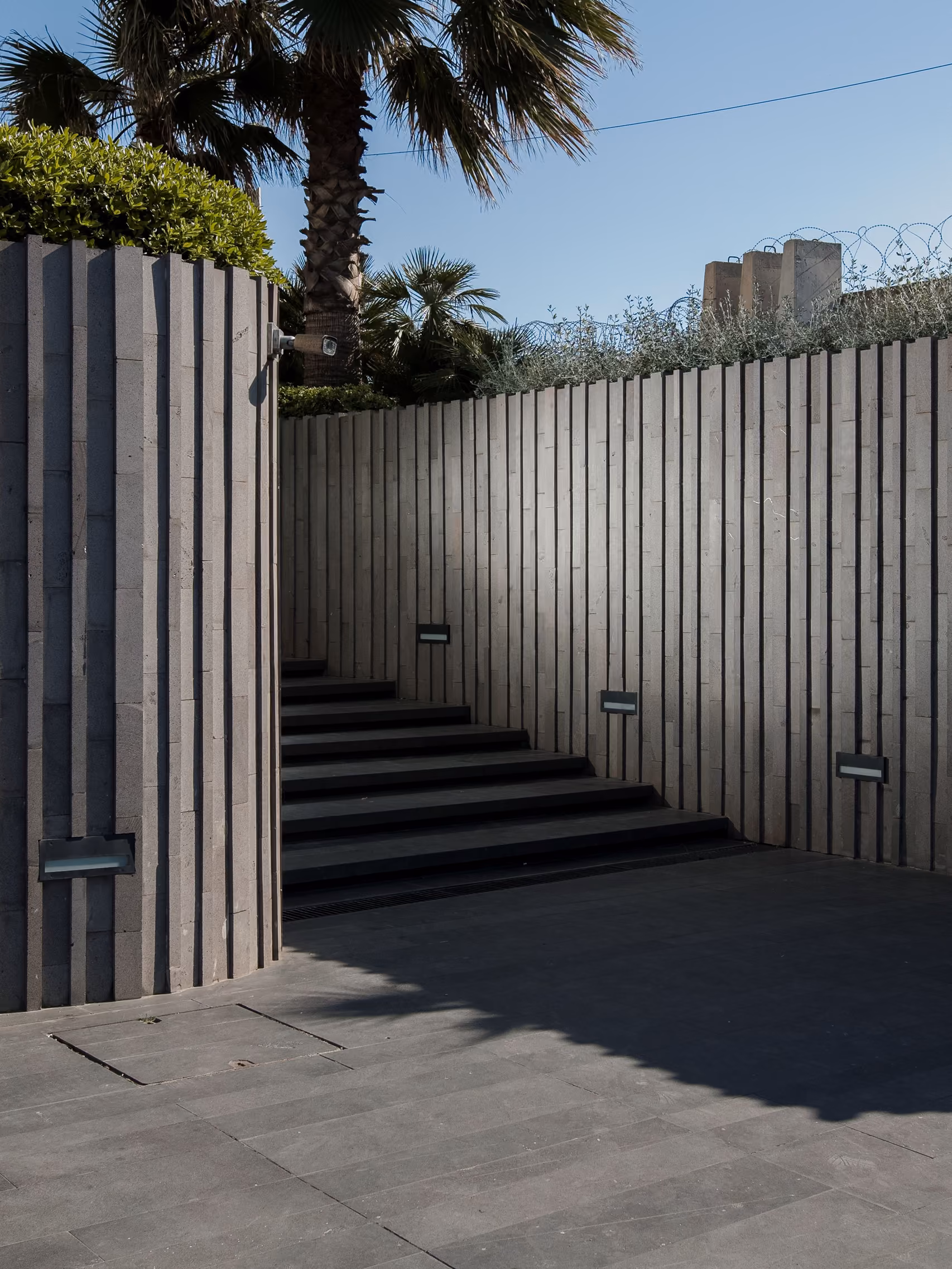 Fluted stone-clad wall at Dalfa Seafront in Beirut, framing a stepped outdoor stair beneath palm trees under a clear sky