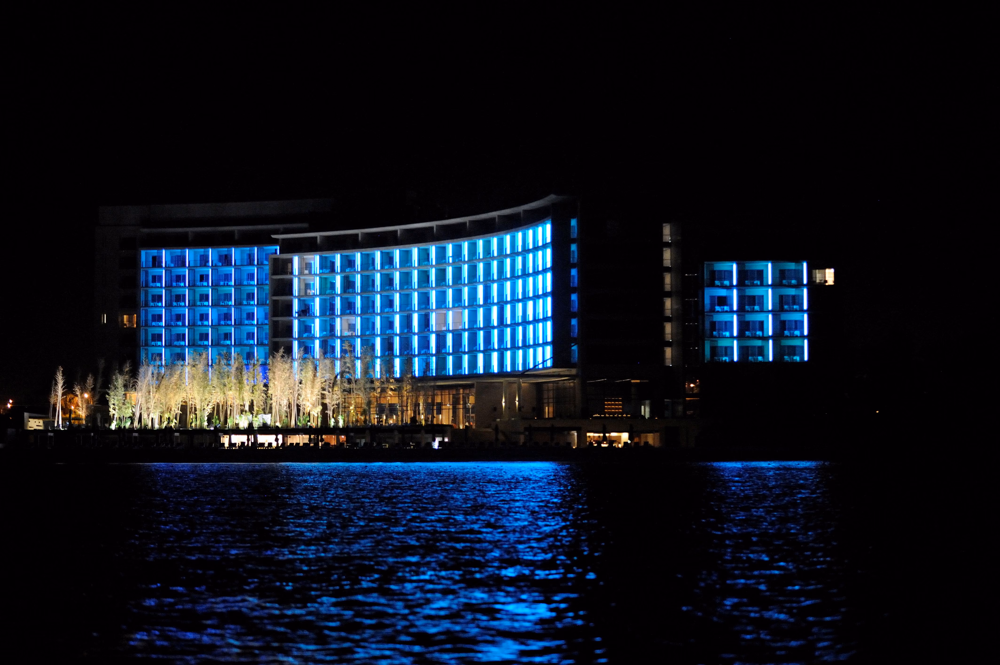 Kempinski Hotel in Aqaba at night, its curved facade glowing blue against the waterfront, seen across dark rippling water