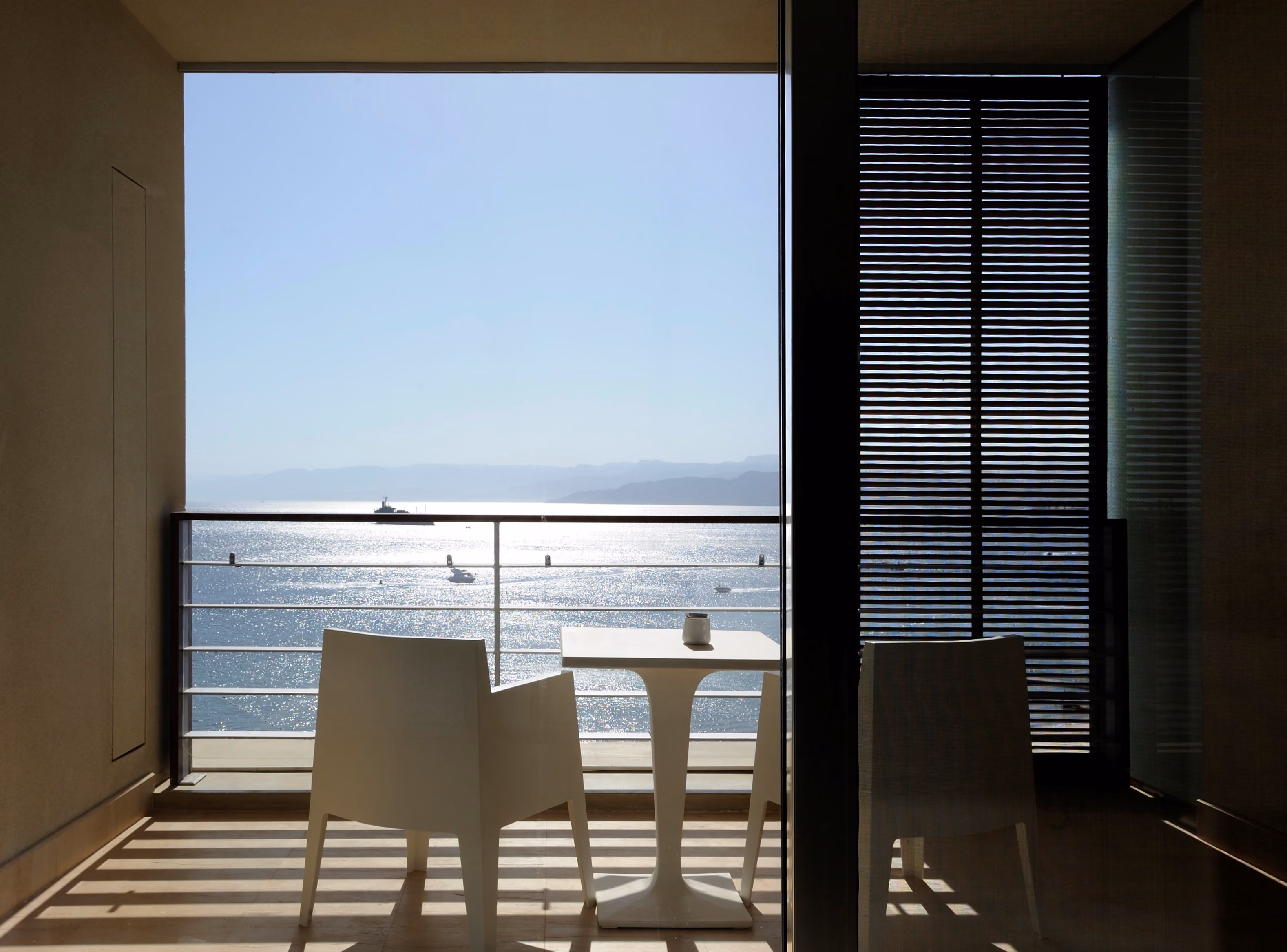 Balcony seating at Kempinski Hotel in Aqaba, white chairs and table beside a glass railing overlooking the sparkling sea and distant mountains