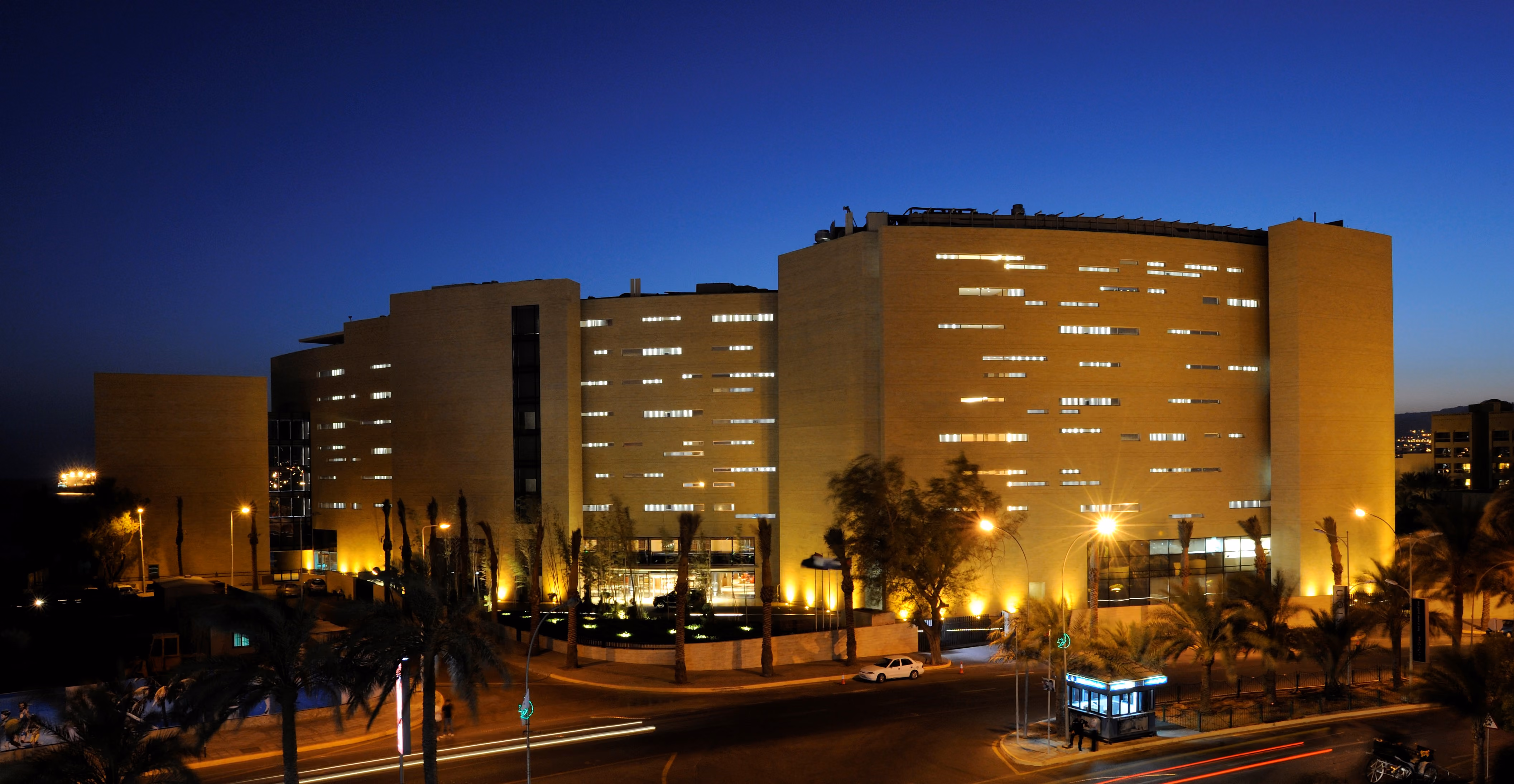 Kempinski Hotel in Aqaba illuminated at dusk, curved yellow stone facade with punctured window openings, palm-lined street in foreground