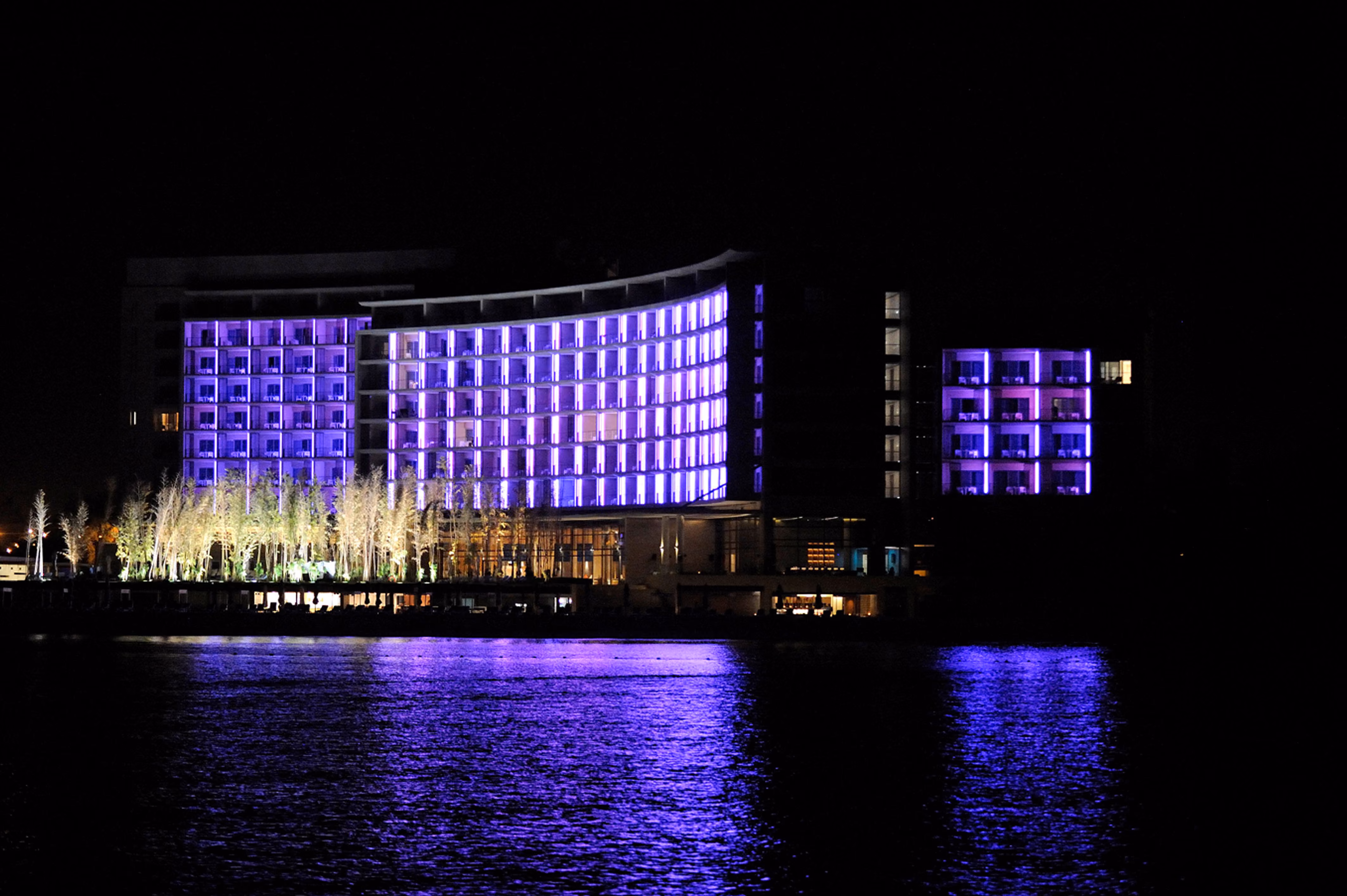 Kempinski Hotel in Aqaba illuminated in purple light at night, curved facade reflecting on the sea with palm trees lining the waterfront