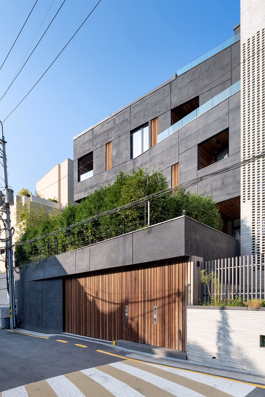 Street view of Pyrite in Seoul, dark textured concrete facade with recessed timber-clad garage and lush greenery spilling over a mid-level terrace