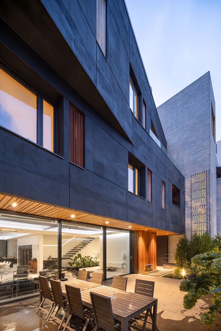Twilight courtyard of Pyrite in Seoul, dark chiselled concrete facade with wood-lined entrance, glazed living room, and an outdoor dining table