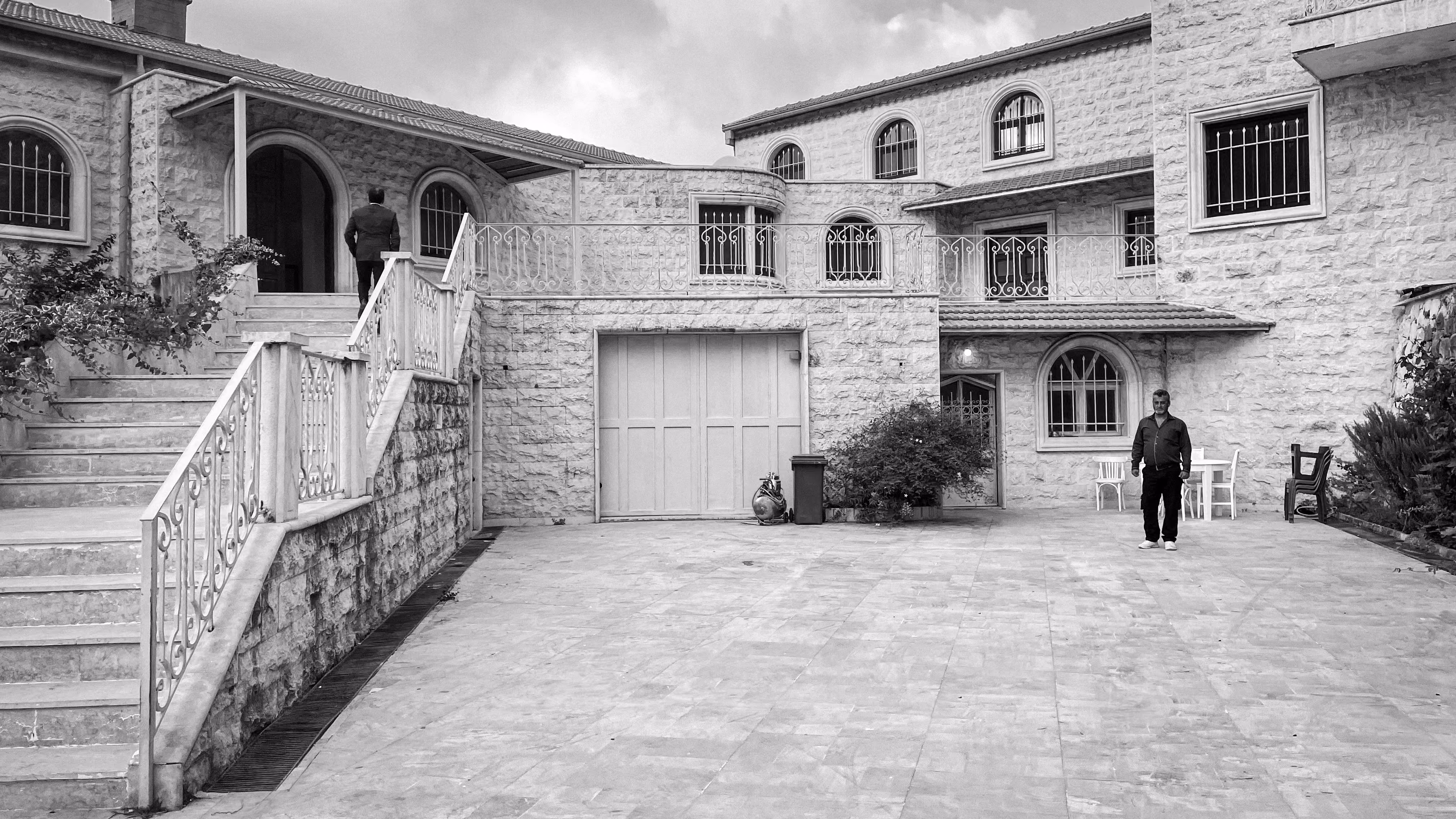 Black and white photograph of YM House in Deir el Qamar, stone courtyard facade with arched windows, wrought-iron balustrades and two men in the paved yard