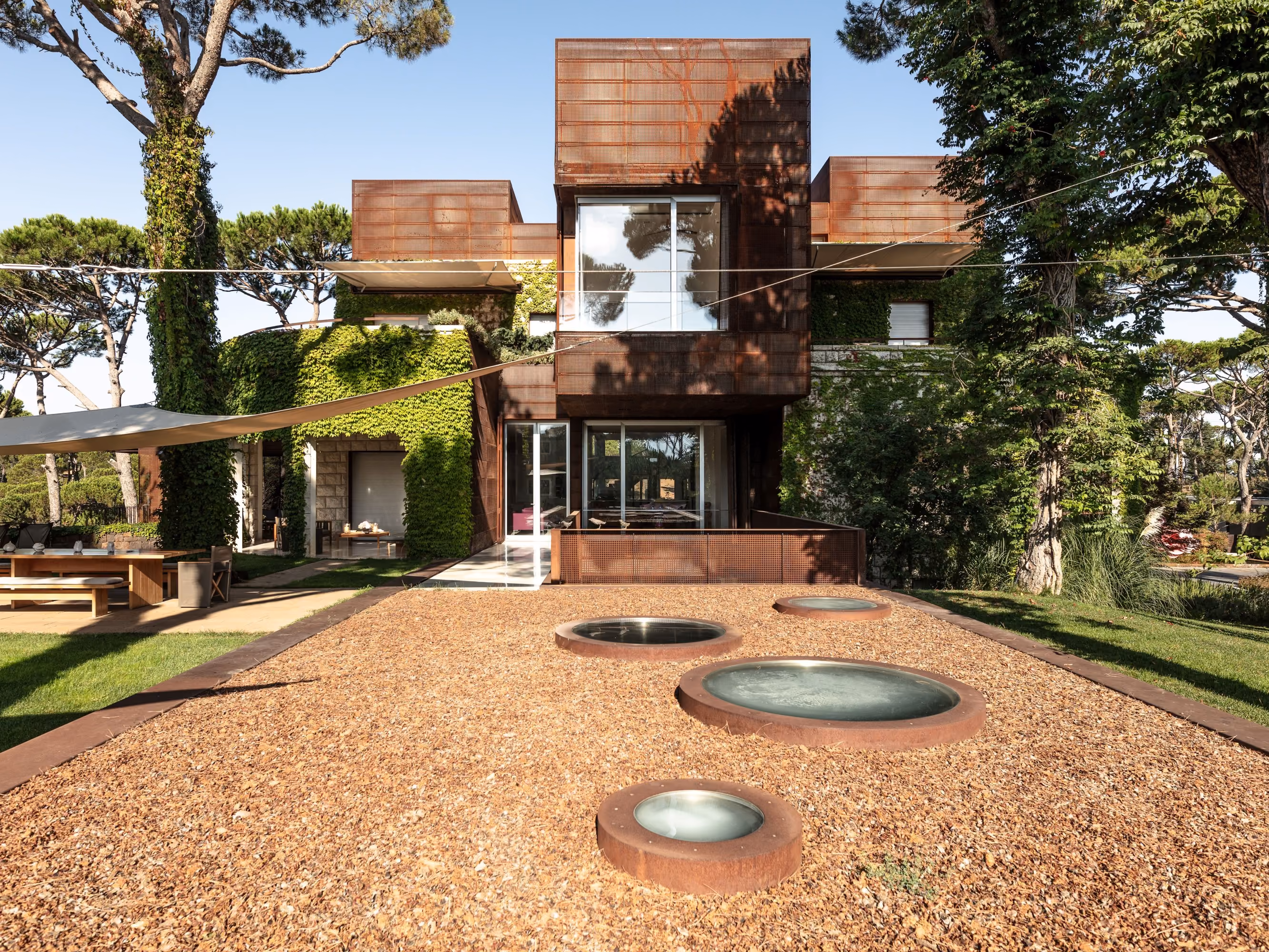 Rear facade of The House with Two Lives in Bois de Boulogne, Corten steel volumes atop a stone base wrapped in climbing ivy, with circular skylights set into a gravel courtyard