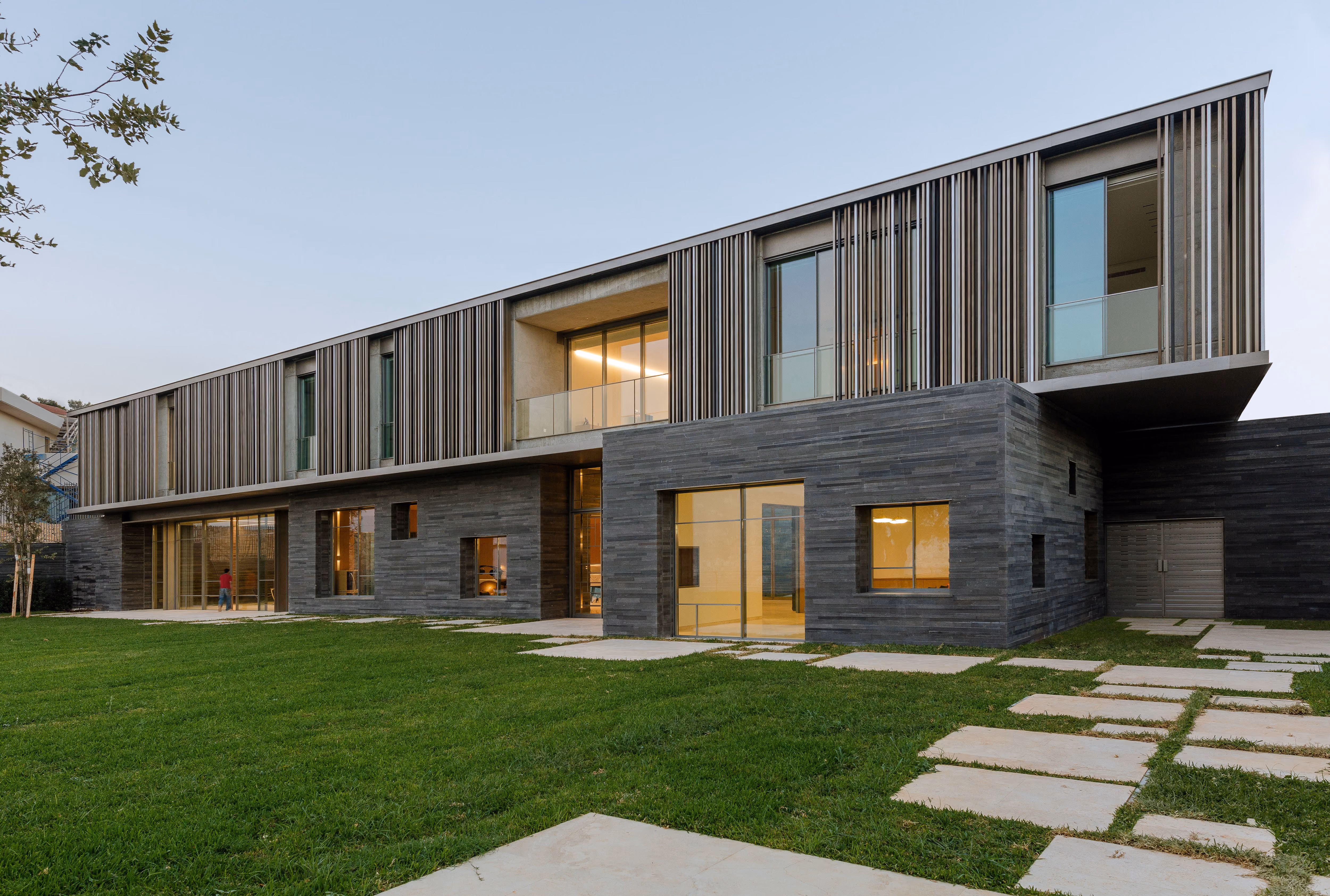 AY House in Yarze, dark stone-clad ground floor with a cantilevered upper level wrapped in vertical timber louvers, overlooking a lawn with stepping stones