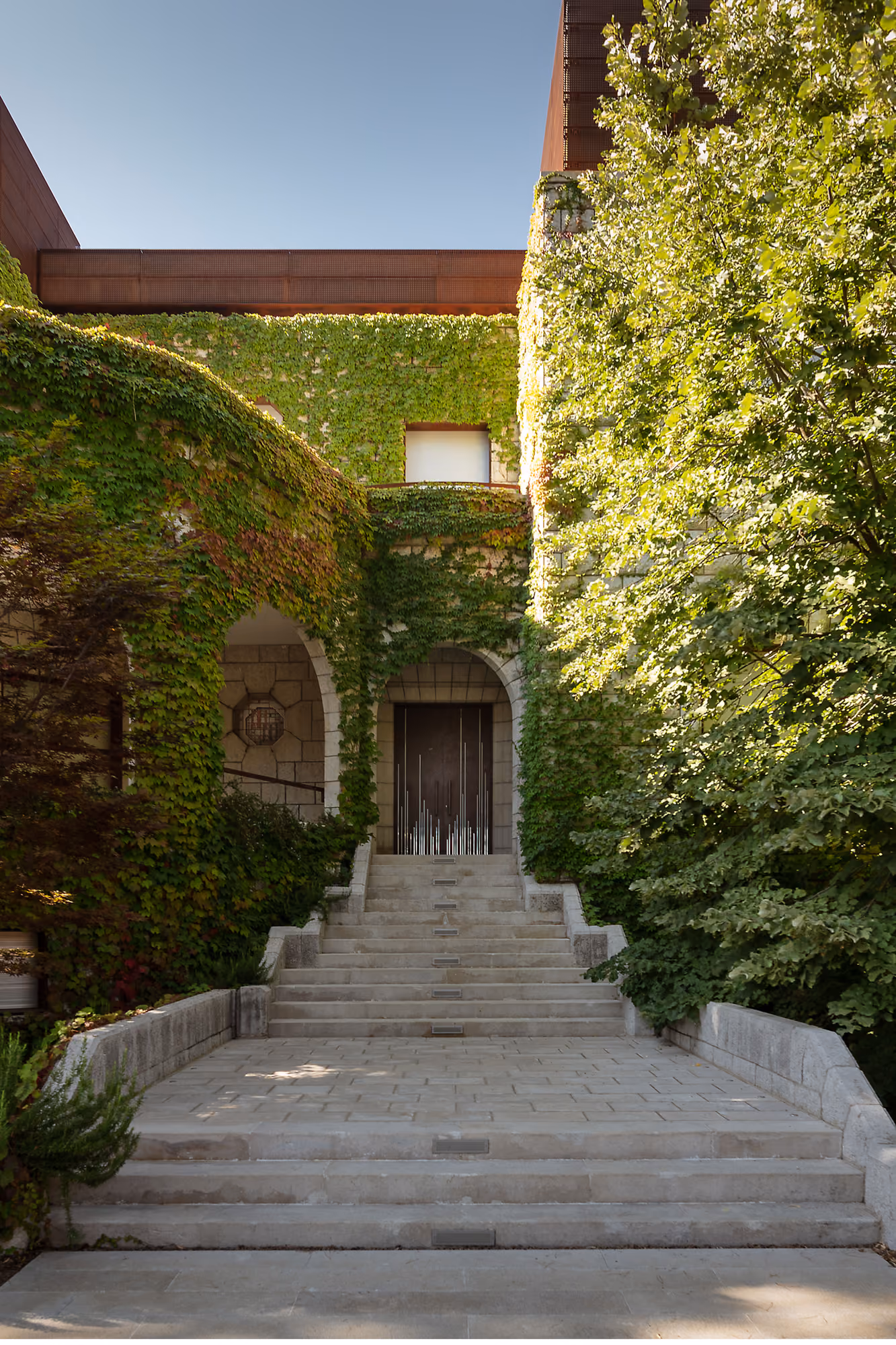 Ivy-covered stone facade of The House with Two Lives in Bois de Boulogne, stone steps leading to an arched entrance beneath a Corten steel roofline