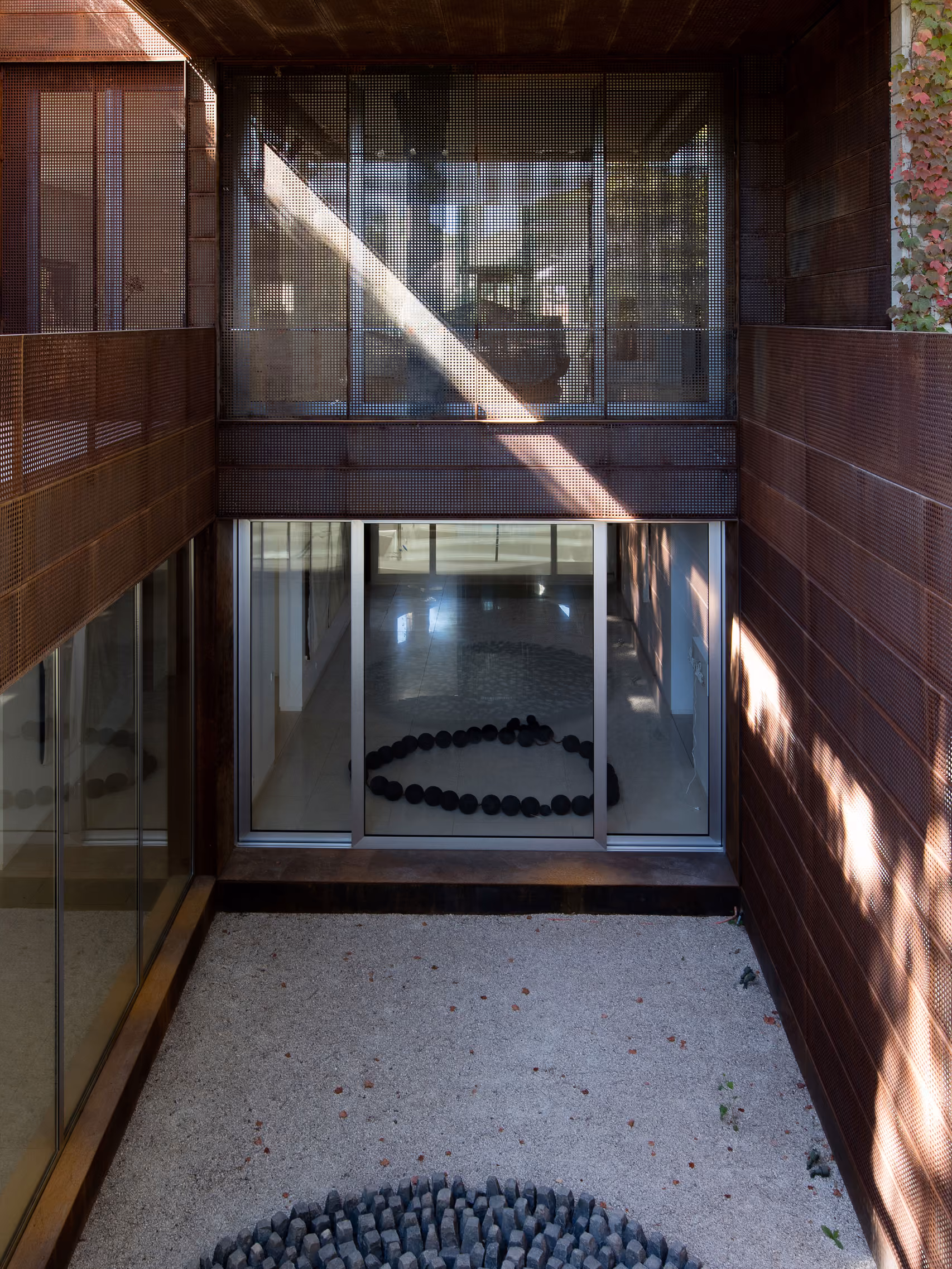 Courtyard view of The House with Two Lives in Bois de Boulogne, perforated Corten steel walls framing glazed openings onto a gravel garden with a circular stone sculpture