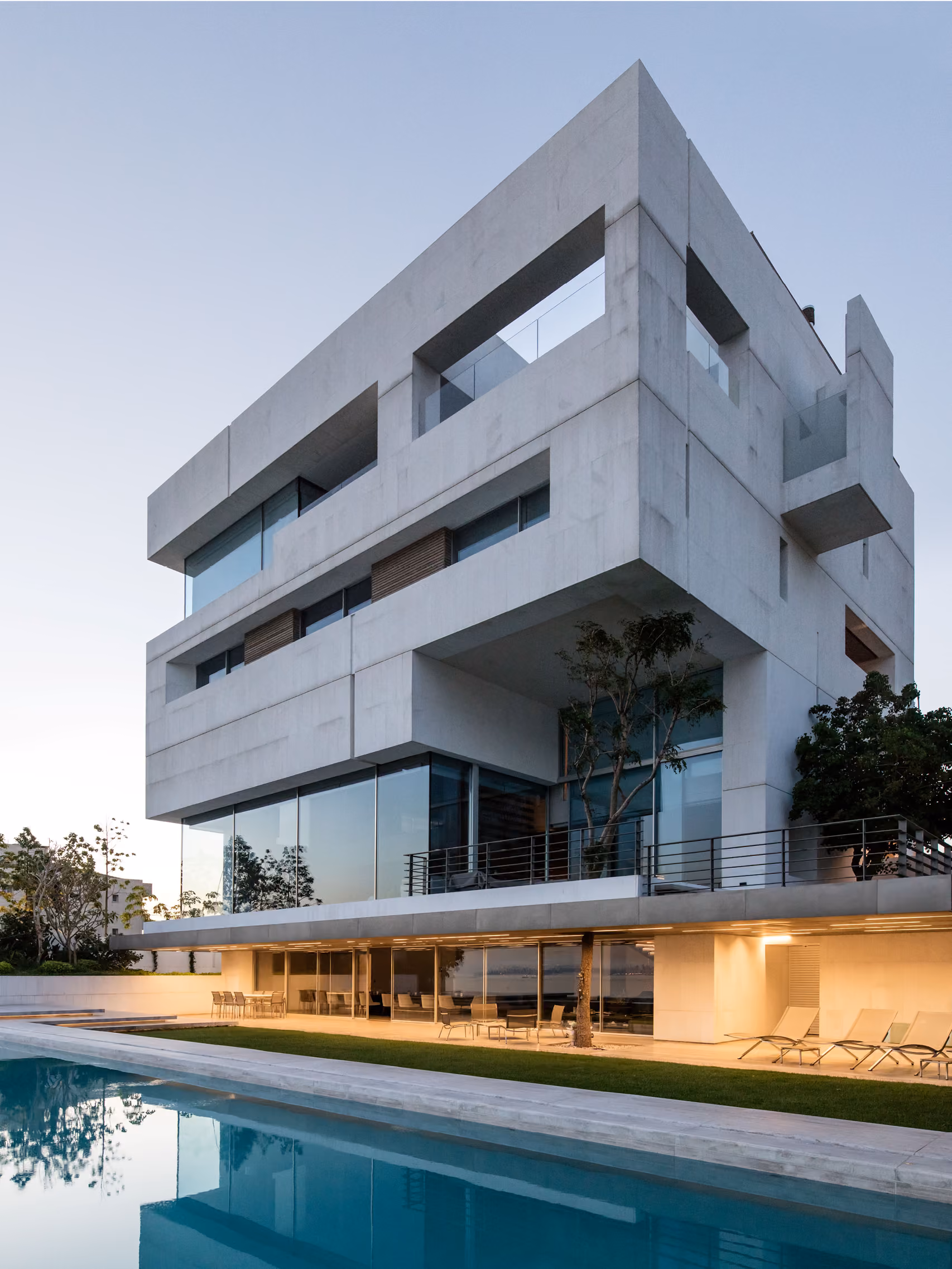 AZ House in Adma, layered concrete volumes with cantilevered cutouts overlooking a garden, glazed ground floor, and lit poolside terrace at dusk