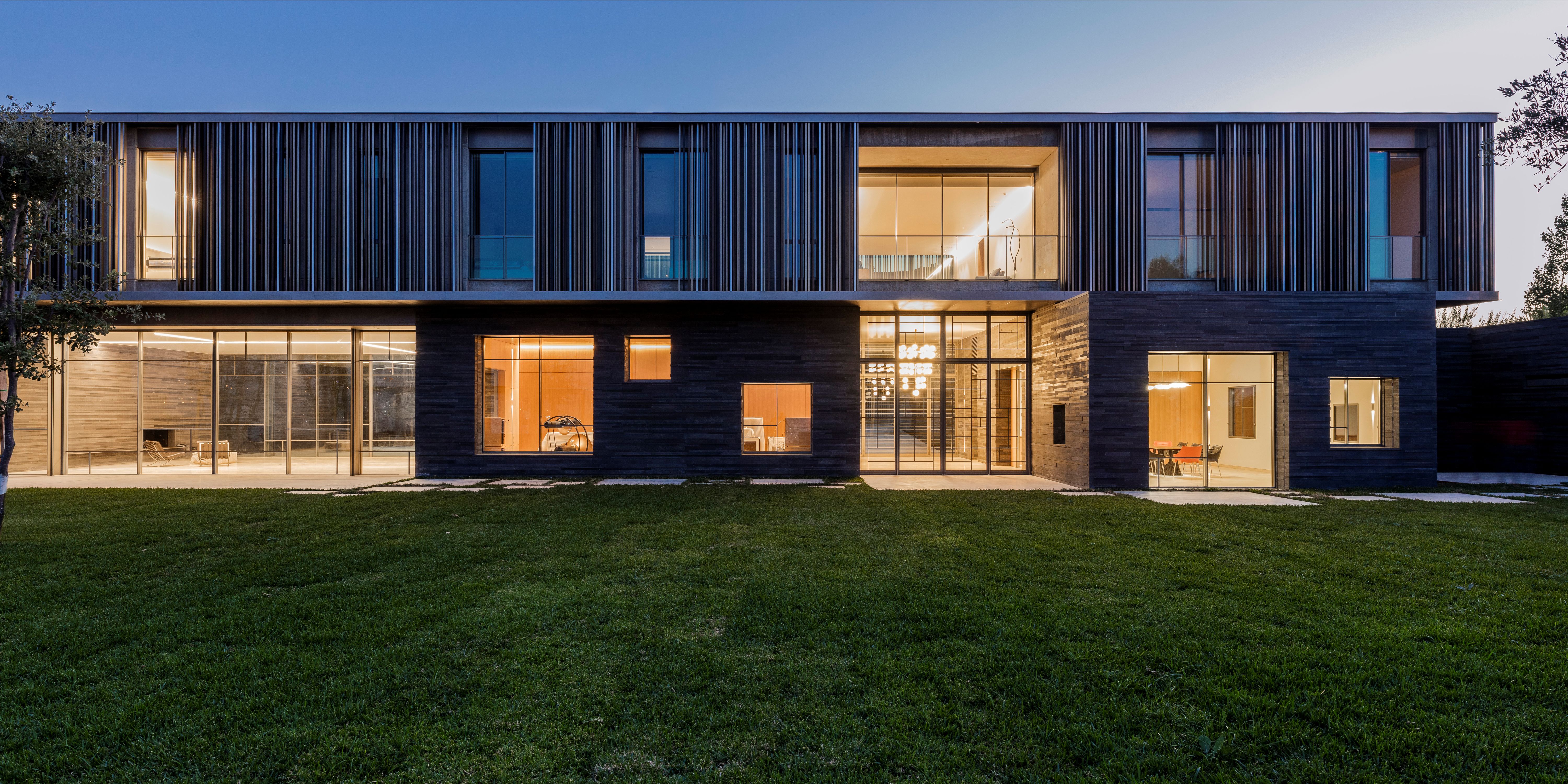 AY House by Nabil Gholam Architects in Yarze, Lebanon, a twilight view of the timber-louvered upper floor cantilevering over glazed ground-floor rooms and a lawn