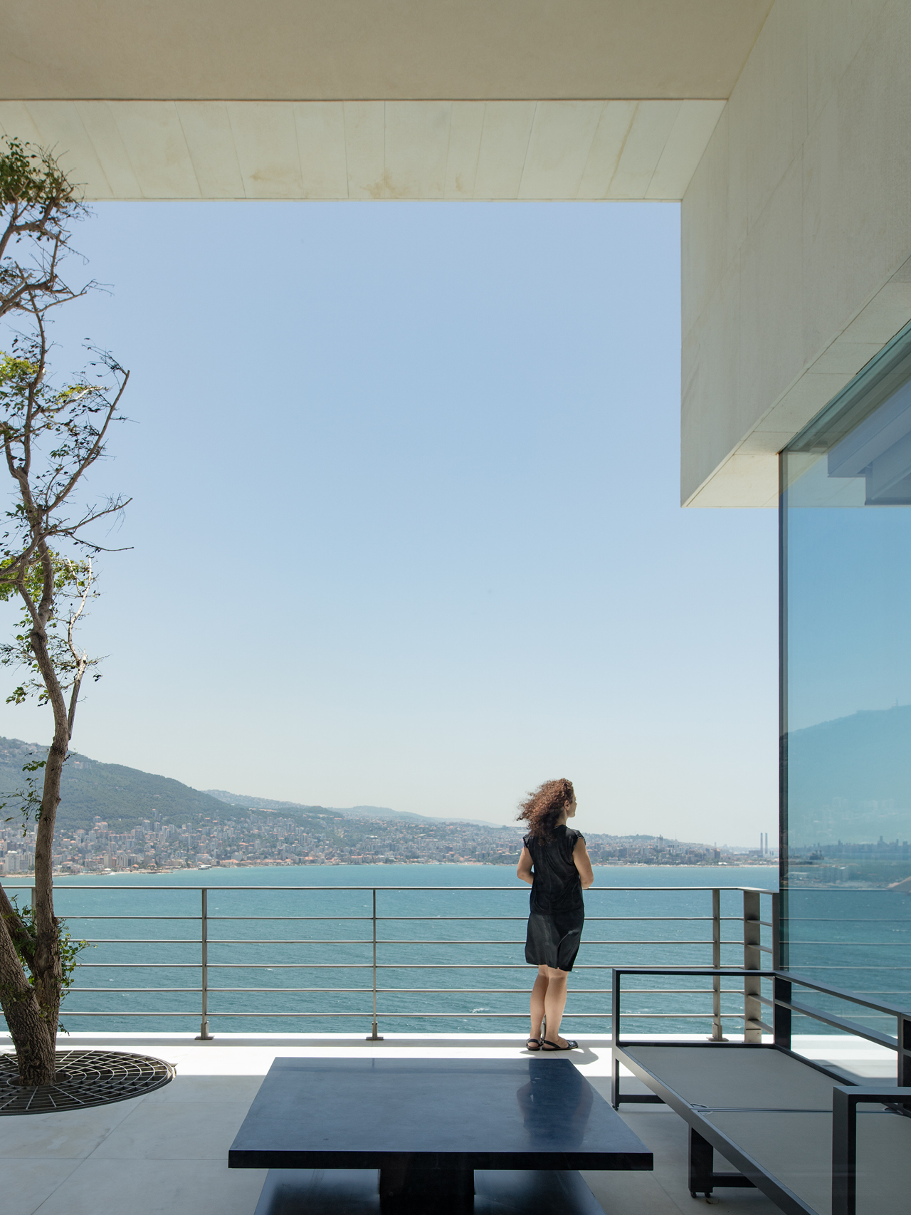 Cantilevered terrace of AZ House in Adma, a figure overlooking the coastline and bay under a concrete roof, framed by glass and a tree in a circular planter