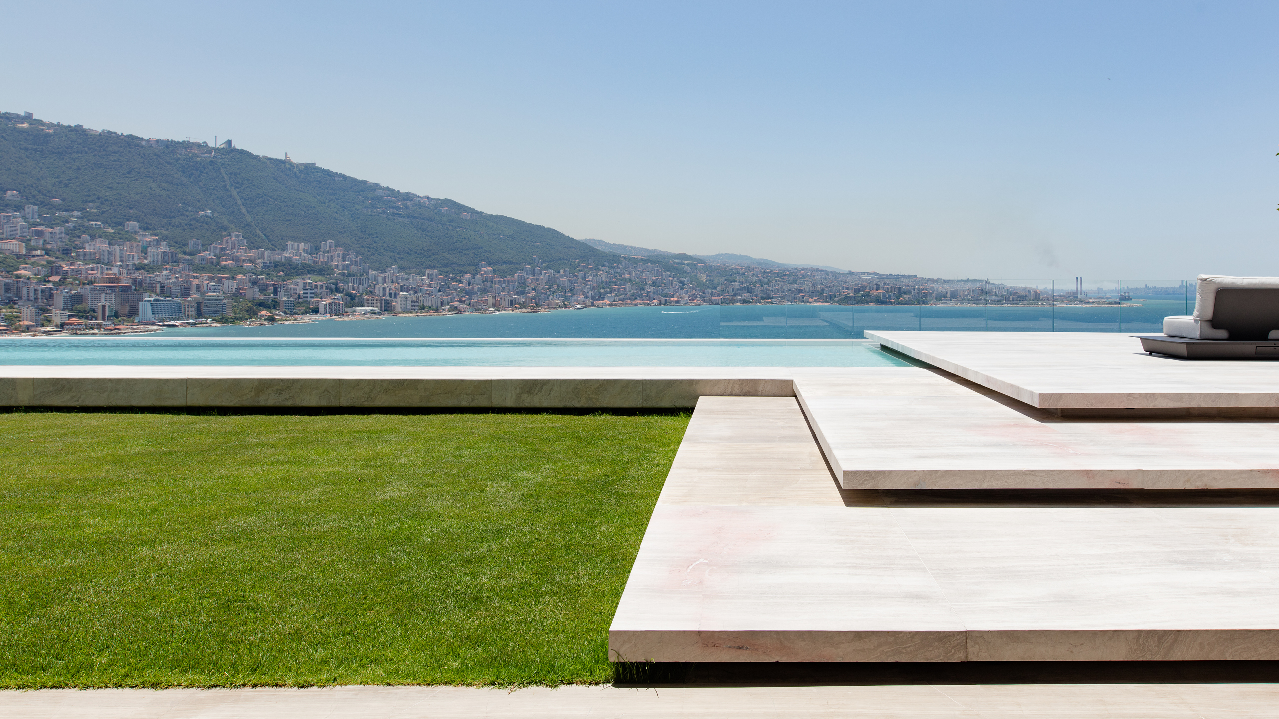 Terraced stone deck and infinity pool at AZ House in Adma, overlooking a lawn, coastal city and mountains along the bay
