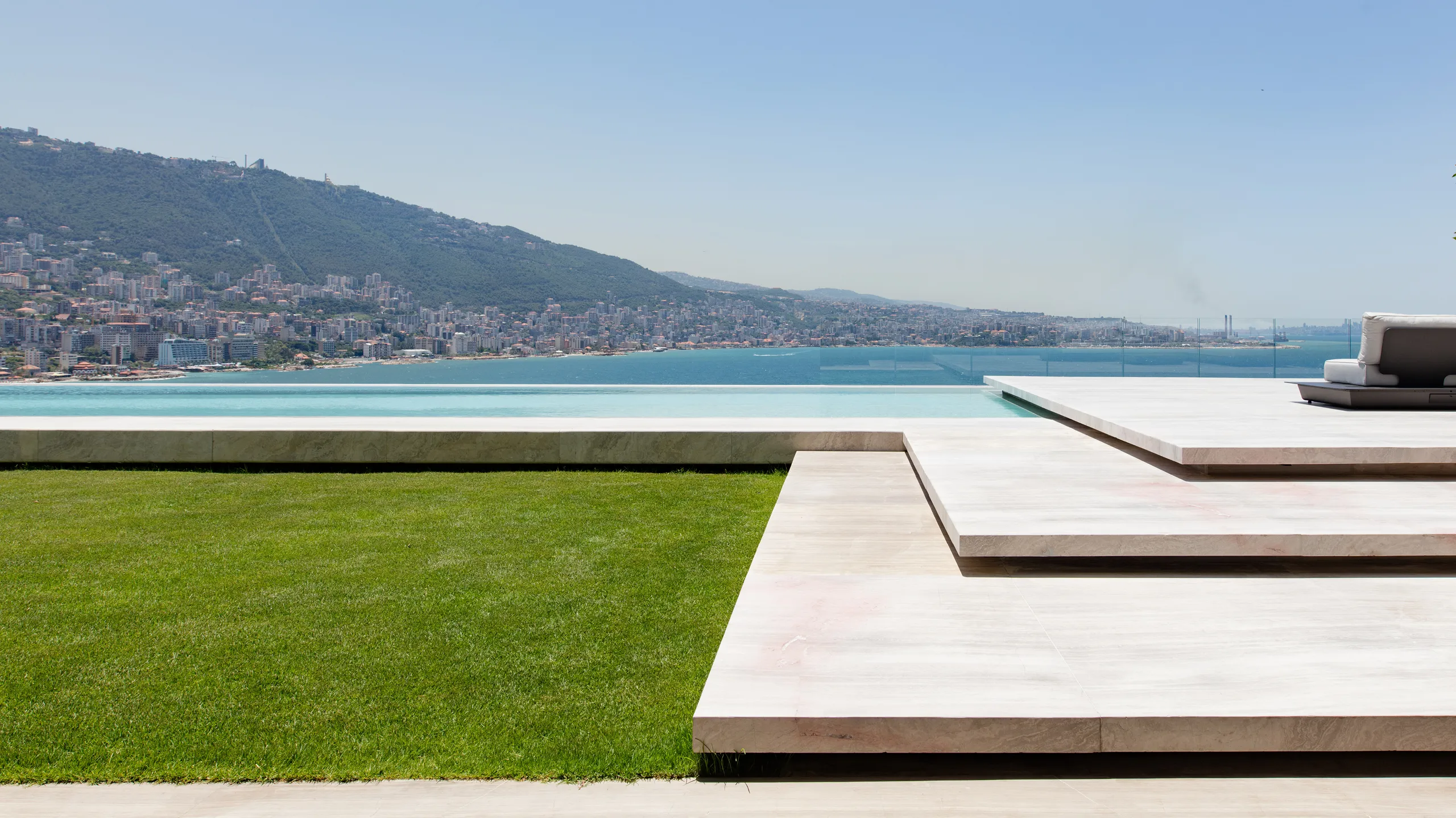 Terraced stone deck and infinity pool at AZ House in Adma, overlooking a lawn, coastal city and mountains along the bay