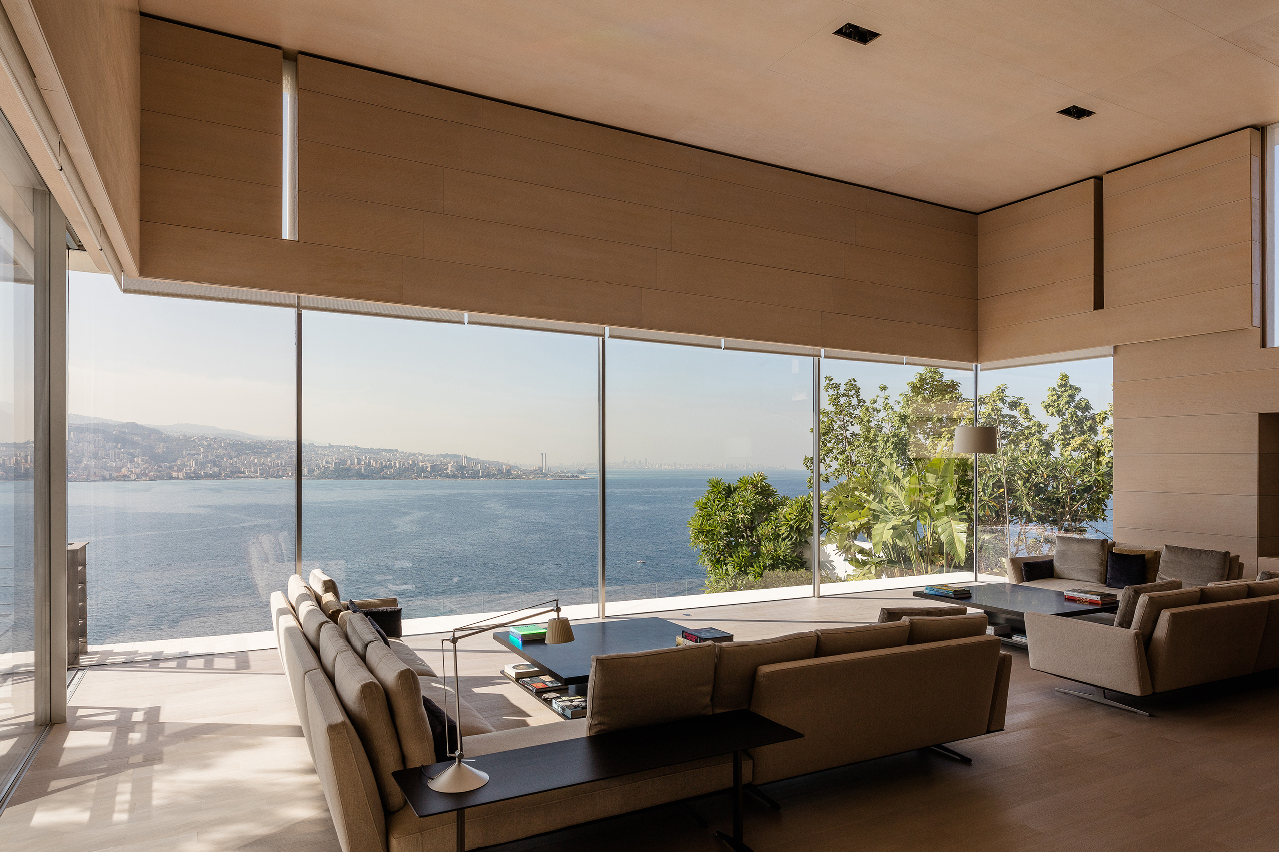 Living room in AZ House, Adma, with wood-clad ceiling and floor-to-ceiling glazing framing sweeping sea and coastline views beyond low sofas