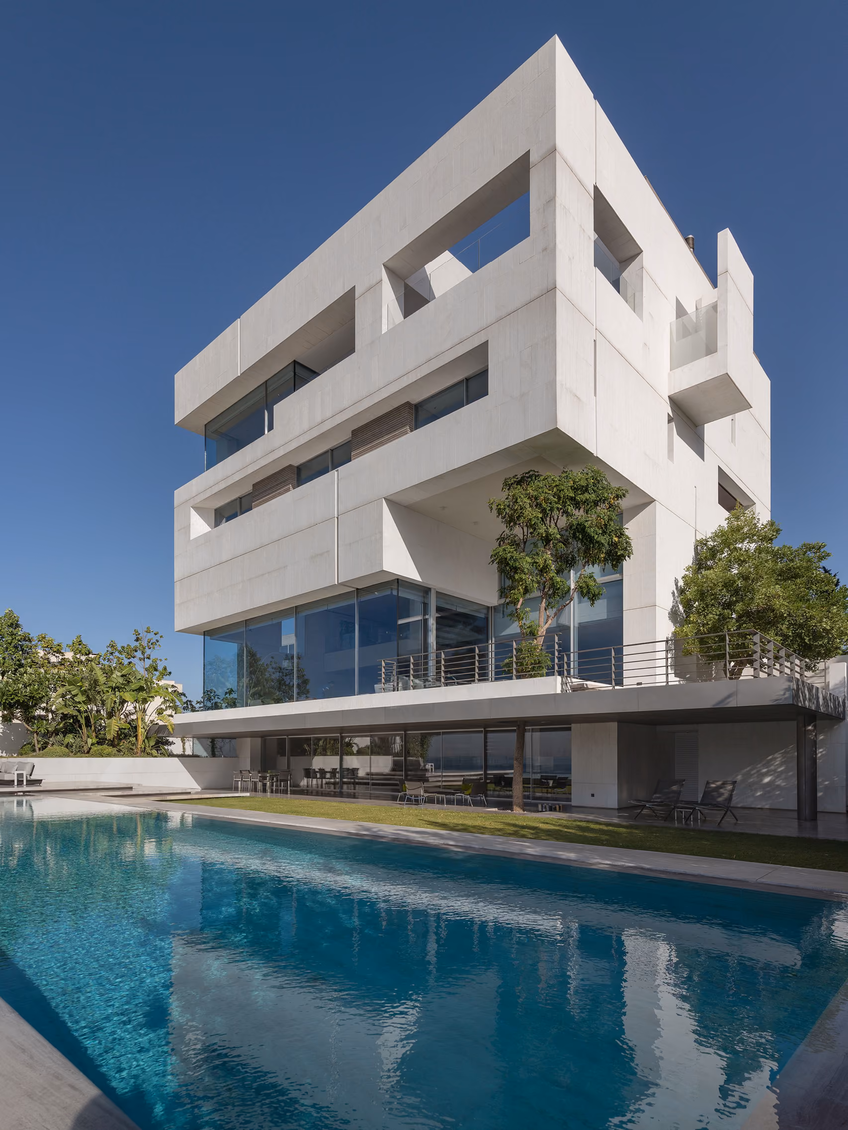 AZ House in Adma, sculptural white cube-like facade with irregular window cutouts, overlooking a long infinity pool and garden terrace