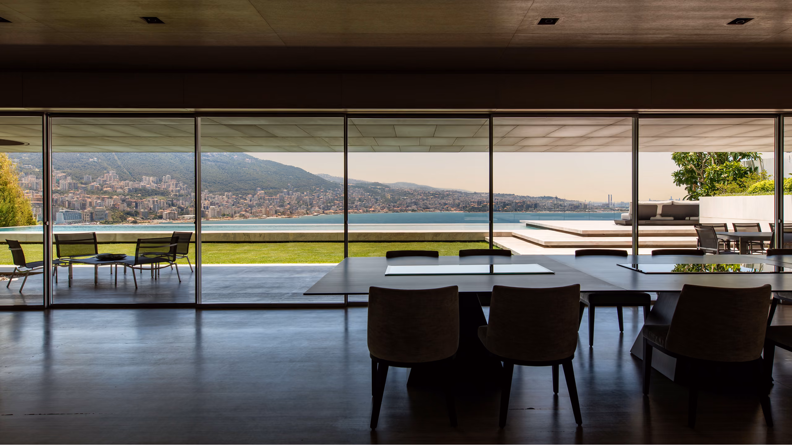 Dining area of AZ House in Adma, glass walls opening to a terrace, lawn and infinity pool overlooking the coastal bay and hills