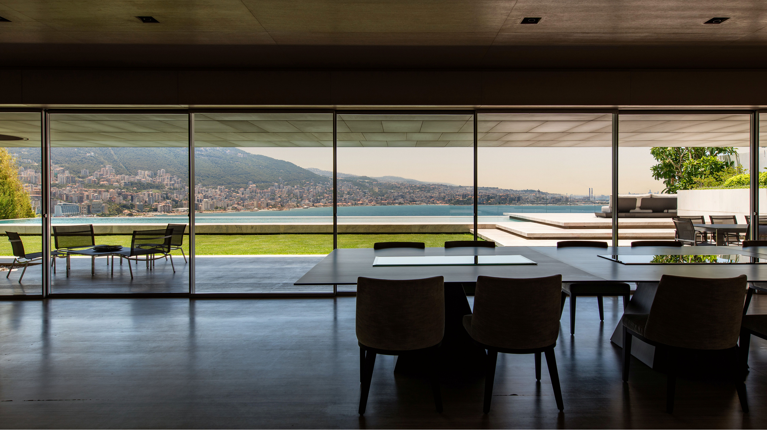 Dining area of AZ House in Adma, glass walls opening to a terrace, lawn and infinity pool overlooking the coastal bay and hills