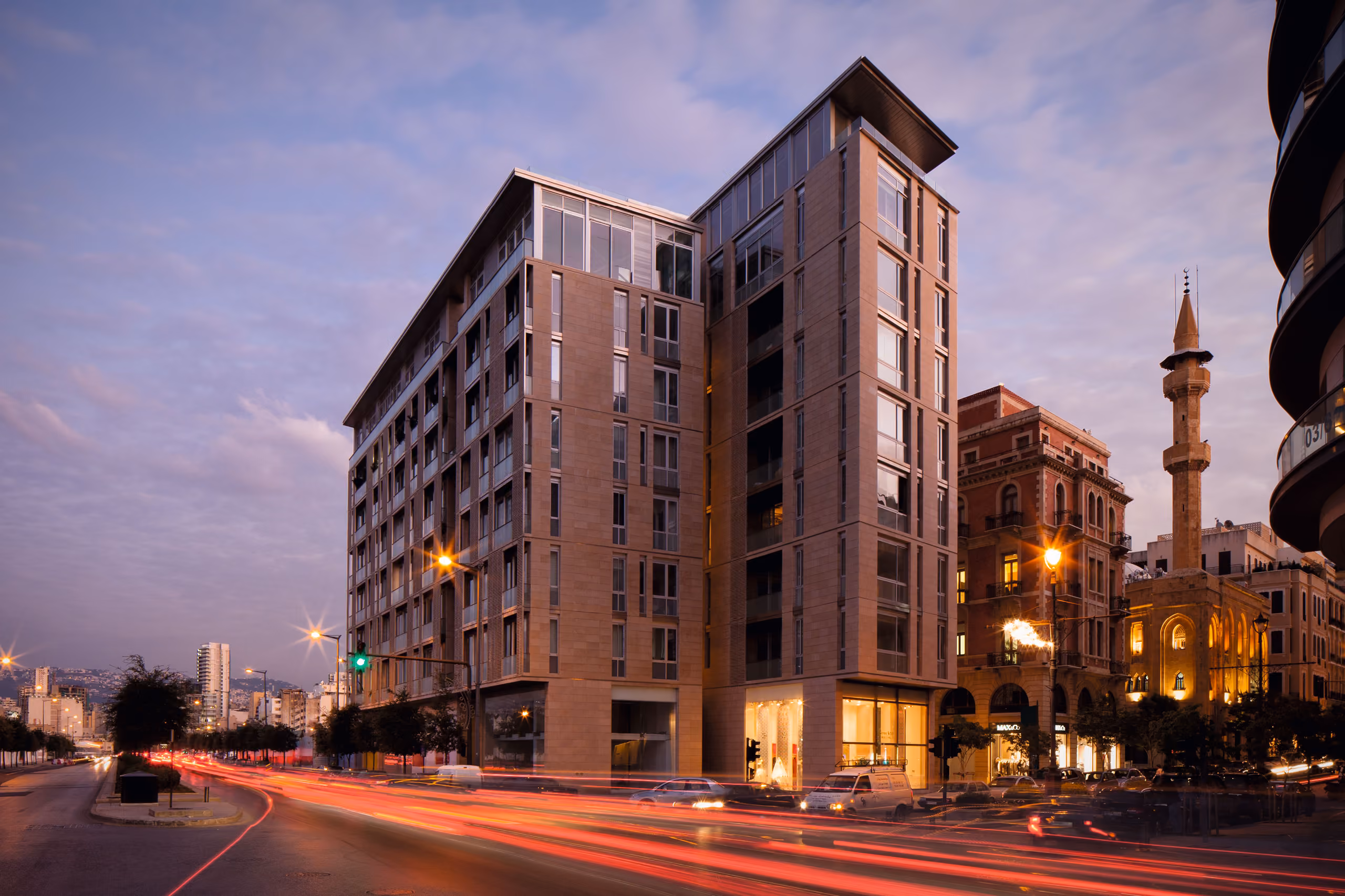 Foch 94 in Beirut Central District at dusk, twin stone-clad residential towers beside a historic mosque, light trails from traffic streaking the street below