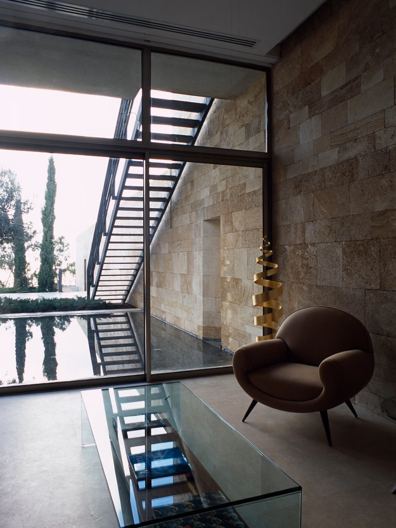 Interior seating area of F House in Dahr el Sawan, stone walls beside a glazed corridor with a floating steel staircase overlooking a reflecting pool