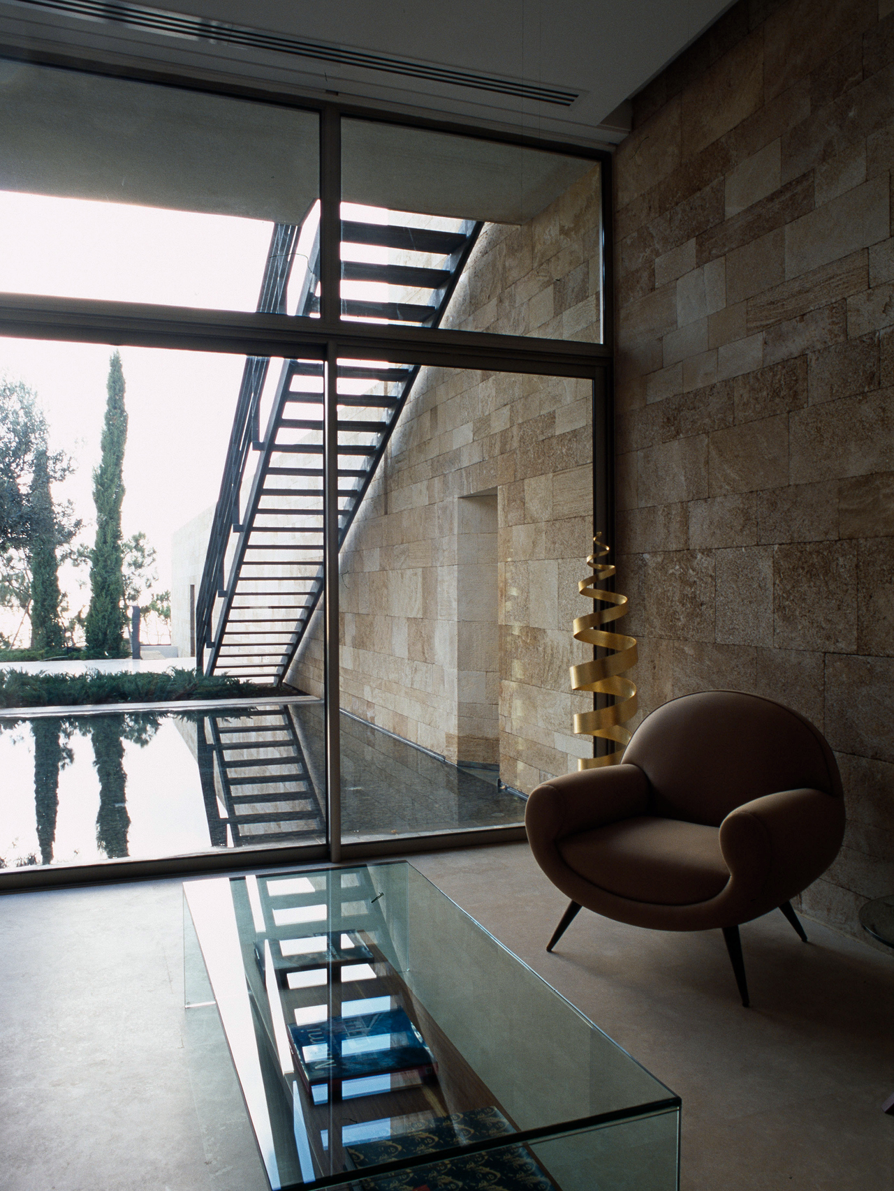 Interior seating area of F House in Dahr el Sawan, stone walls beside a glazed corridor with a floating steel staircase overlooking a reflecting pool