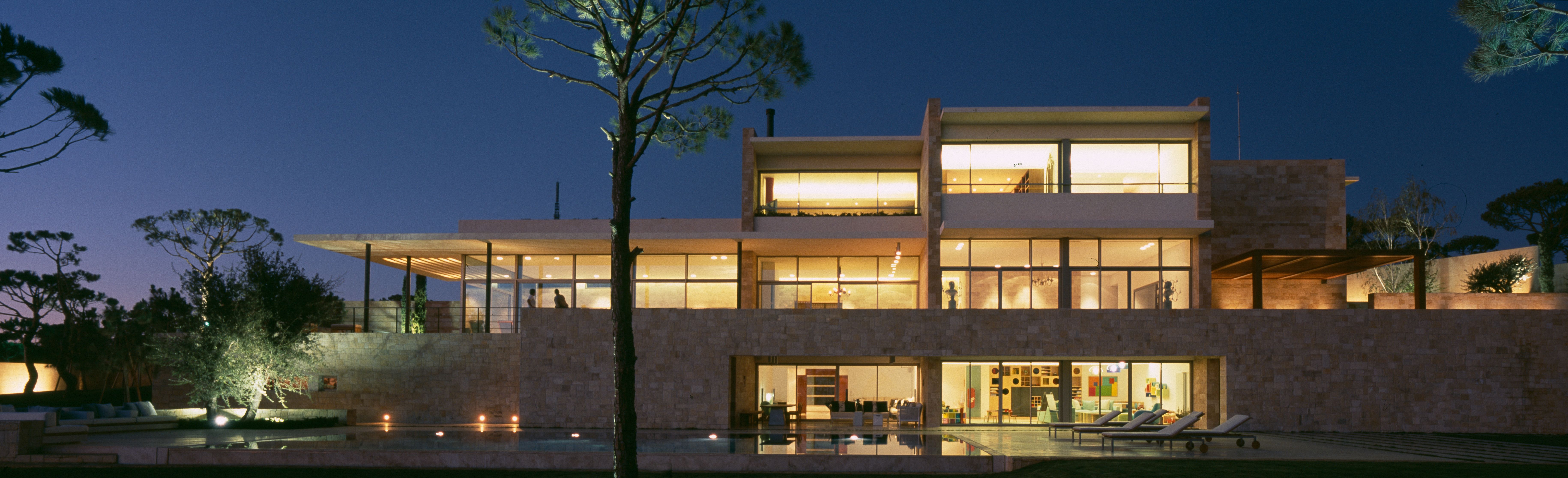 F House by Nabil Gholam Architects, illuminated at dusk in Dahr el Sawan, Lebanon, stone walls and glazed volumes overlooking a reflecting pool and pine trees