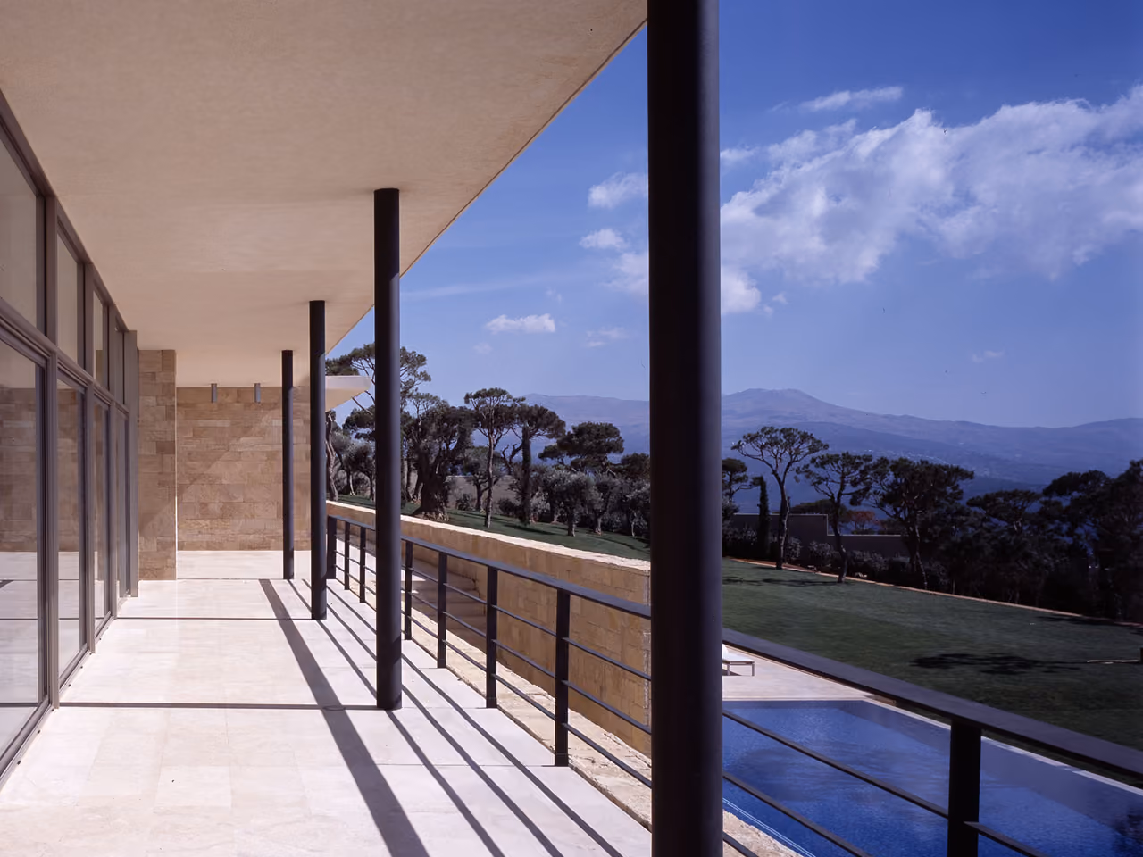 Covered terrace of F House in Dahr el Sawan, stone columns lining a travertine walkway beside a pool, with pine-covered hills stretching to distant mountains