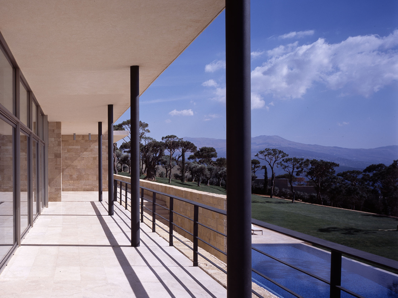 Covered terrace of F House in Dahr el Sawan, stone columns lining a travertine walkway beside a pool, with pine-covered hills stretching to distant mountains
